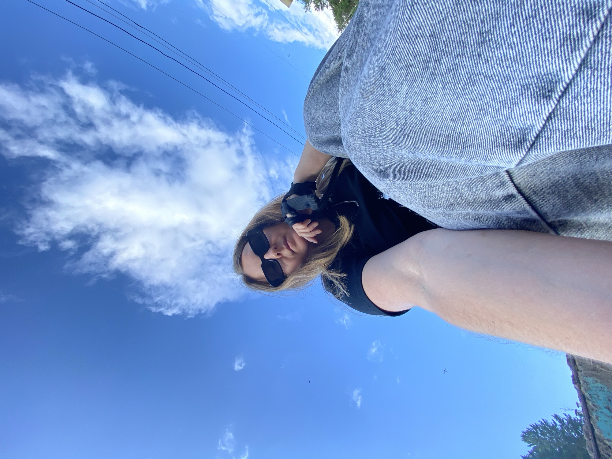 The image shows a low-angle, wide-shot selfie of a person sitting outdoors with a bright blue sky and scattered clouds in the background. The person is wearing sunglasses, a black t-shirt, and grey denim pants. Their left arm is extended towards the camera, and their right hand is resting on their chin. In the distance, a few birds are visible in the sky, and power lines are strung across the upper right portion of the frame. The lower left of the image shows a blurry green and brown textured surface, possibly a wall or railing, with some trees in the background. There is no visible indication of the specific location being Noviy, Russia.