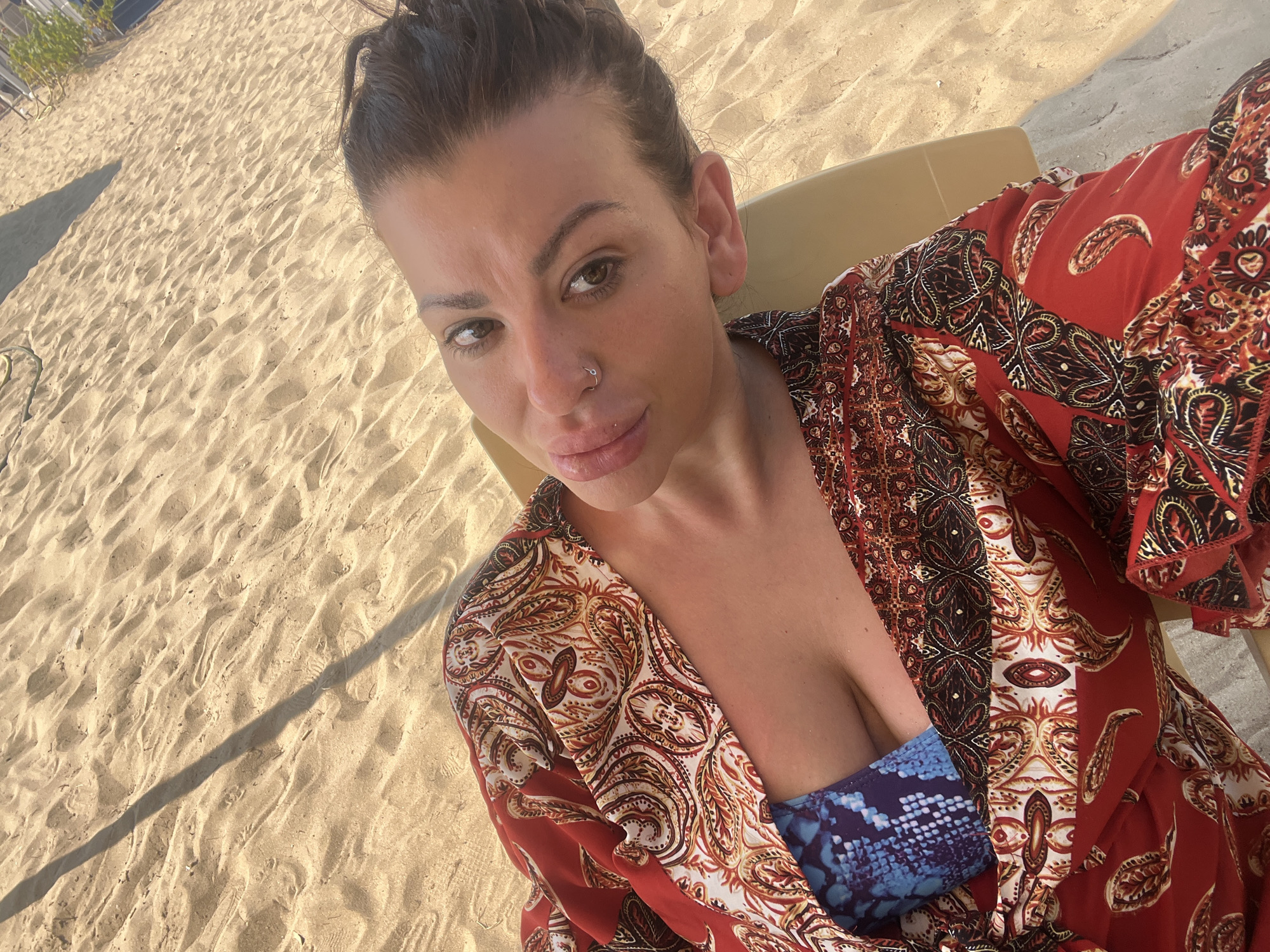 A woman is lying on a beach chair on a sandy beach. She is wearing a red, patterned kimono over a blue and black patterned bikini top. The sun is shining and casting a shadow across the sand. The background shows more sand and a glimpse of some vegetation and a building. The woman is looking at the camera with a slight smile. The location is likely a beach in Sozopol, Bulgaria, given the typical scenery of the area.