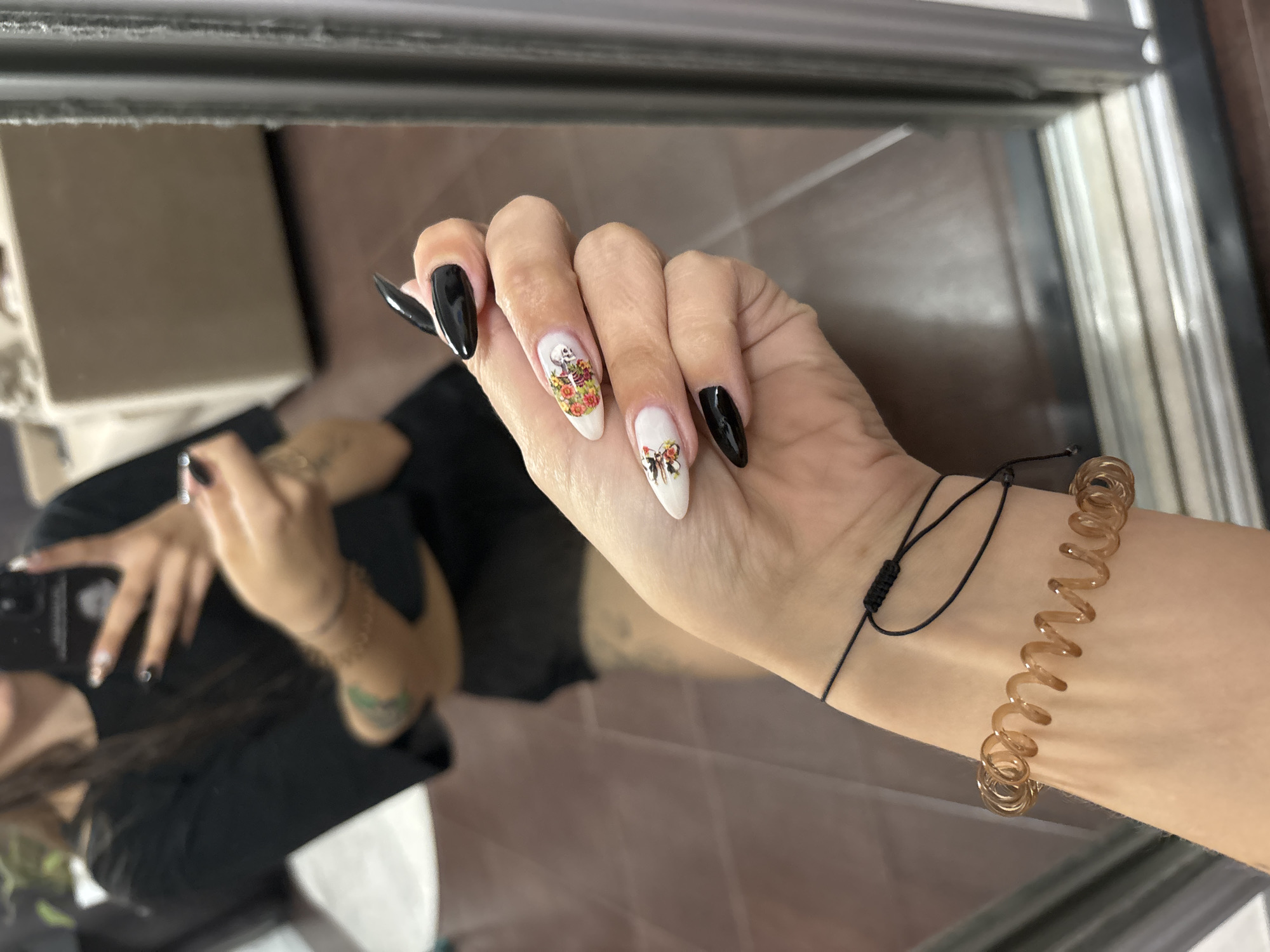 The image shows a close-up of a person's hand and arm, which is reflected in a mirror. The person has long, stiletto-shaped fingernails, with some painted black and others decorated with white polish and decals. One decal appears to be a skeleton surrounded by flowers, and another shows a bird with flowers. The person is wearing a black t-shirt and has tattoos on their arm. A thin black string bracelet is tied around their wrist, and a spiral-shaped bracelet is worn below it. The reflection shows the person holding a phone, presumably taking the picture, and a glimpse of furniture in the background. The flooring appears to be made of brown tiles. The image does not contain any information that would allow for the determination of its location in Novokuznetsk, Russia.