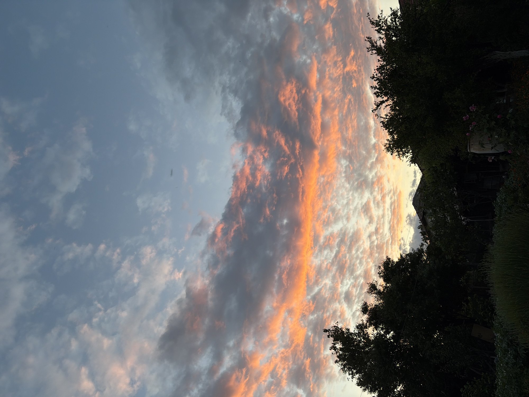 The image depicts a sky with streaks of orange and gray clouds, likely at sunset or sunrise. To the right, there is a silhouette of trees and a structure, possibly a house or building, partially obscured by foliage. The location is a city named Unknown in a region also named Unknown, but specific details about the area are not discernable.