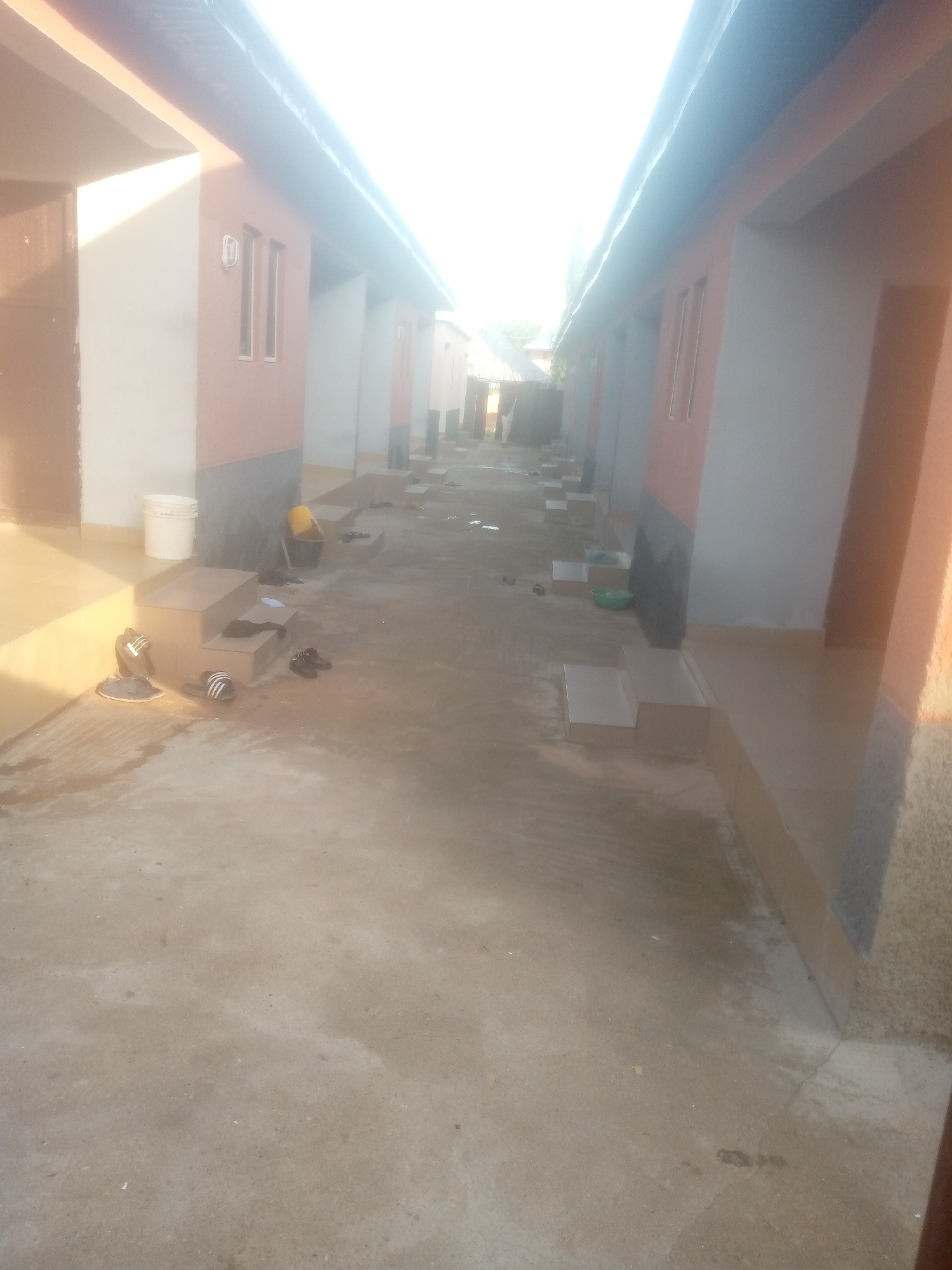 This is a photograph of a row of buildings with pale peach and gray facades in Jalingo, Nigeria. The buildings appear to be single-story units with small windows and doors. A concrete walkway runs in front of the buildings, and there are several pairs of sandals and flip-flops scattered on the ground and on a set of steps leading up to one of the units. A white plastic bucket and a yellow container are also visible near the steps. The ground is a light brown concrete, and the scene is illuminated by bright sunlight, creating strong shadows. The overall impression is one of a residential or commercial area, possibly a low-income housing complex or shopfronts.