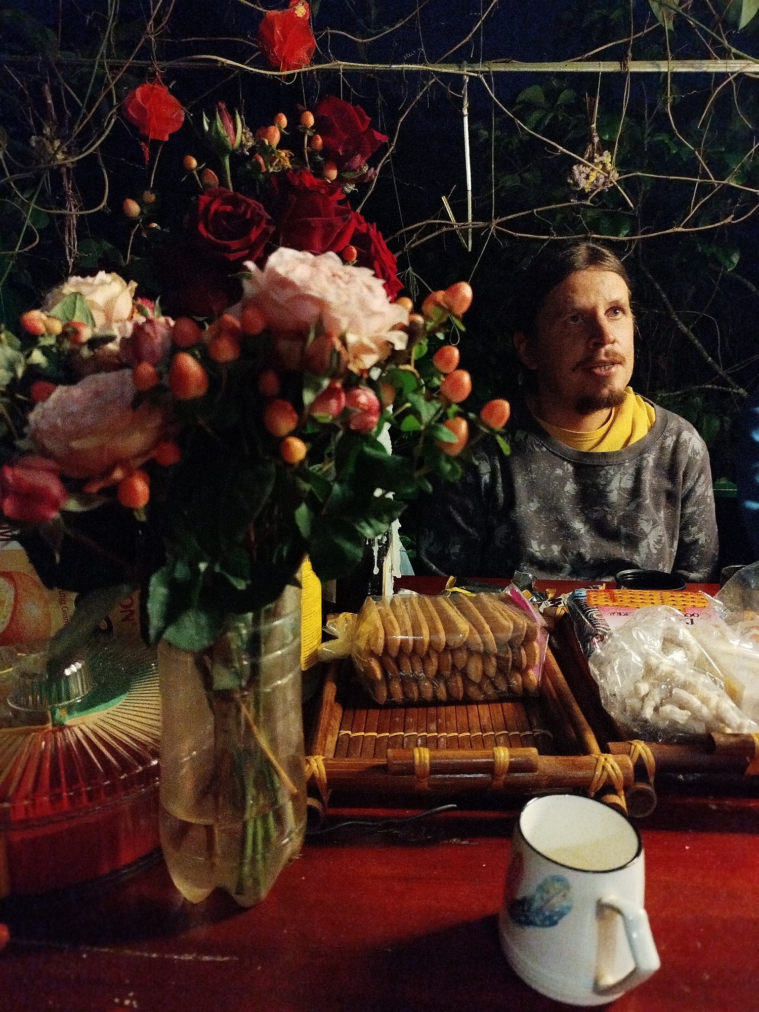 The image depicts a person sitting at a table laden with items, with a bouquet of roses as a prominent feature. The setting appears to be outdoors, possibly a garden or patio, given the presence of foliage and branches in the background.
The individual at the table has medium-length hair, a mustache, and a beard. They are wearing a gray patterned sweater over a yellow shirt. Their gaze is directed slightly upwards and to the right.
On the table, a vase made from a plastic bottle holds a bouquet of roses in shades of pink and red, along with green foliage and small orange berries. Other objects on the table include a packaged stack of cookies, a package of white snacks, a red container, and a white mug with a blue design. The surface of the table is red.
Based on the provided information, the location is Cam Ly - Da Lat, Vietnam. The scene suggests a casual gathering or a moment of relaxation in an outdoor setting.