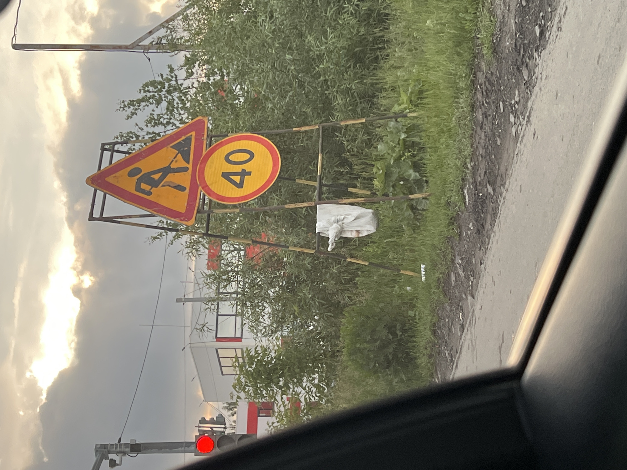 The image depicts a street scene in Yekaterinburg, Russia, with a focus on a traffic light and road signs. A traffic light with a red illuminated signal is visible on the left side of the frame. To the right of the traffic light, a yellow triangular road sign with a red border indicates road work, showing a silhouetted figure digging with a shovel. Below this, a circular yellow sign with a red border displays the number "40," signifying a speed limit of 40 kilometers per hour. These signs are mounted on a metal structure. A white cloth object is hanging from the structure. The background features a building with white walls and red accents, along with abundant green vegetation and trees. The sky is overcast with sunlight breaking through the clouds. The foreground shows a portion of a road and what appears to be the interior of a vehicle, suggesting the photo was taken from a car.