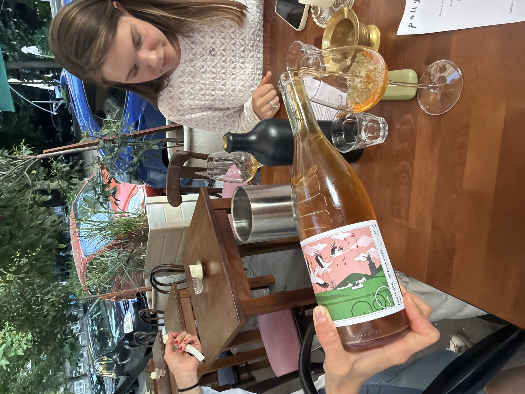 An overhead shot captures a group of people dining outdoors at a table. The scene is set in Belgrade, Serbia, indicated by the restaurant's menu in the foreground. A woman with long brown hair, wearing a white textured top, is seated on the right, looking down at the table. In the center, a person's hand is pouring amber liquid from a green and pink labeled bottle into a wine glass. The bottle has text that reads "MALVASIA - Skin contact 2024." Other objects on the table include a black bottle, a metal trash can, a gold-colored holder, and a smartphone. In the background, parked cars and lush green trees frame the outdoor dining area.