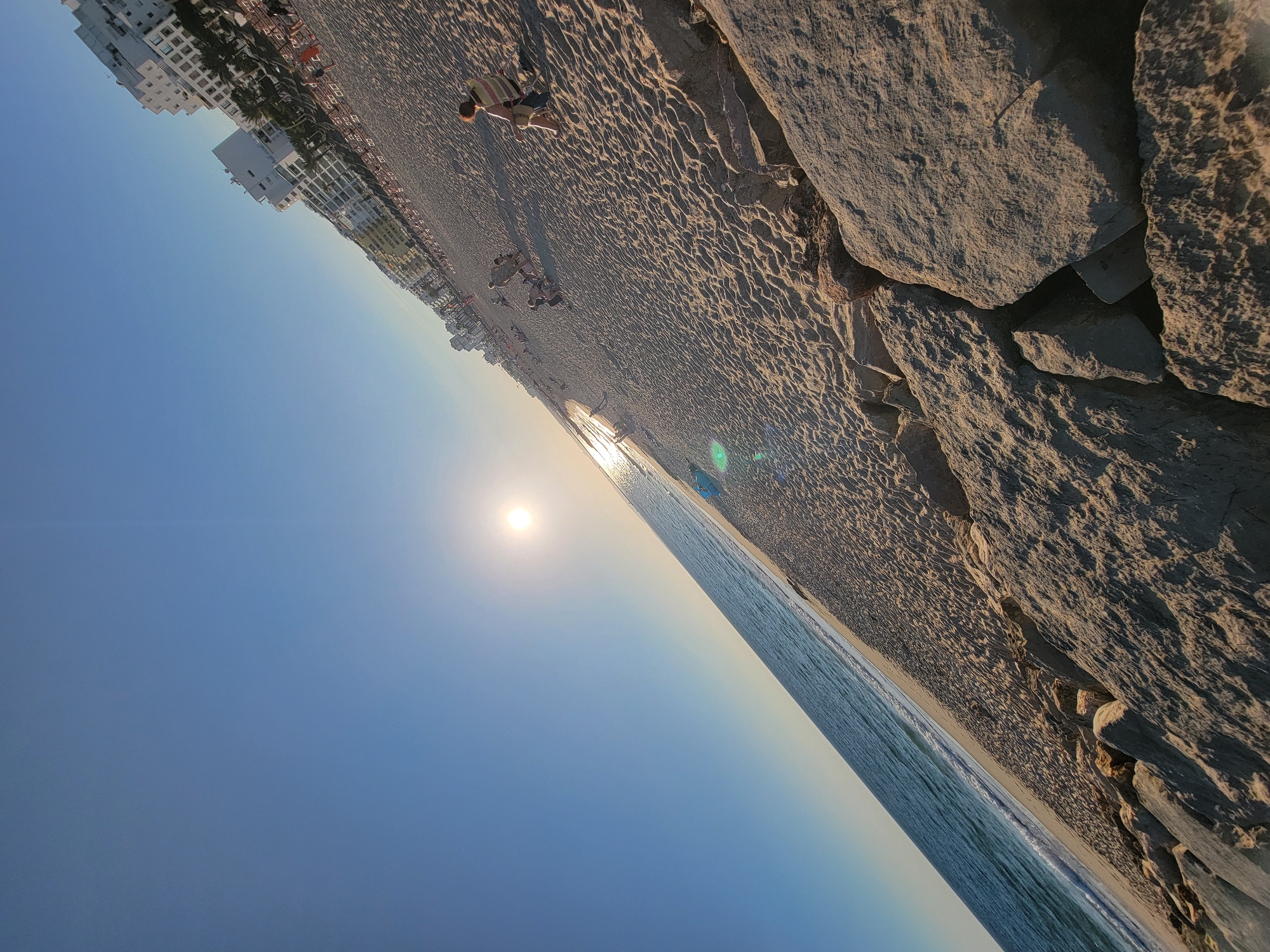 The image shows a wide, sandy beach in Quarteira, Portugal, during what appears to be late afternoon or early evening, given the low angle of the sun. The sky is a clear, bright blue, with the sun positioned in the upper left quadrant, casting a bright glare. The ocean is a calm, deep blue with gentle waves lapping at the shore. The beach is populated with people, some relaxing on lounge chairs, others walking or playing in the sand.
Along the coastline, a row of white, multi-story buildings with many windows lines the edge of the beach, indicating a developed resort area. The sand is packed and textured, with visible footprints and ripples. In the foreground, large, grey rocks are piled along the edge of the beach, possibly forming a breakwater or barrier. The perspective of the image is tilted, with the horizon line diagonally bisecting the frame. A few individuals are visible on the sand, including a person sitting in a striped chair and a child with brown hair. Beach umbrellas and lounge chairs are arranged in rows further down the beach.