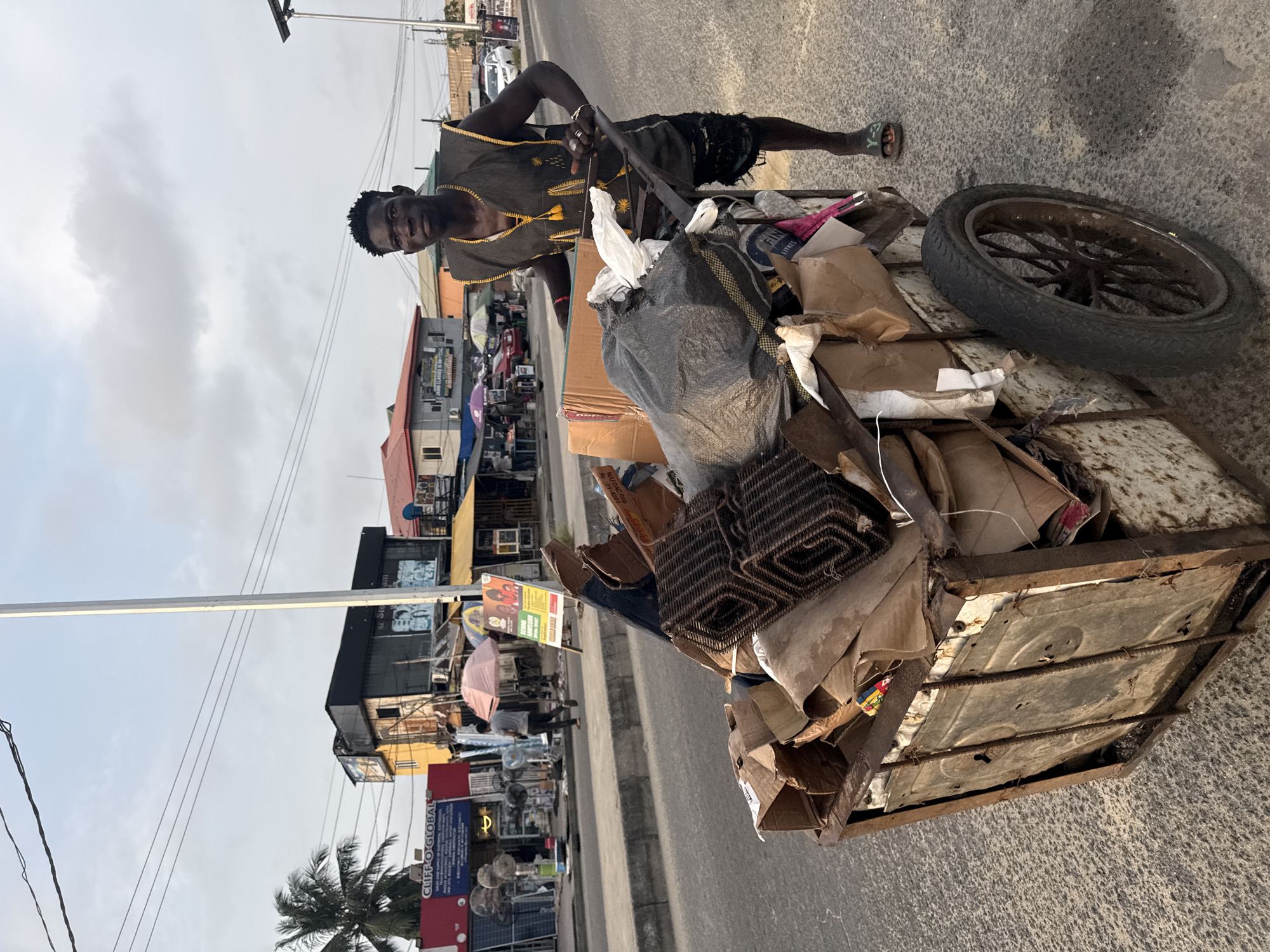 A young Black male, wearing a dark sleeveless vest with yellow trim and trim shorts, stands next to a dilapidated cart filled with assorted debris on a paved street in Ado, Nigeria. The cart, made of rusted metal, contains cardboard boxes, plastic bags, and what appear to be rolled-up mats or rugs. The man is gripping the handle of the cart, suggesting he is about to move it or has just stopped. Behind him, a street scene unfolds with various buildings, businesses with signage, and a few other individuals in the distance. A palm tree is visible on the left side of the frame. The sky above is partly cloudy, with overhead wires crisscrossing the scene. The overall impression is of a bustling urban environment where individuals engage in scavenging or waste collection.