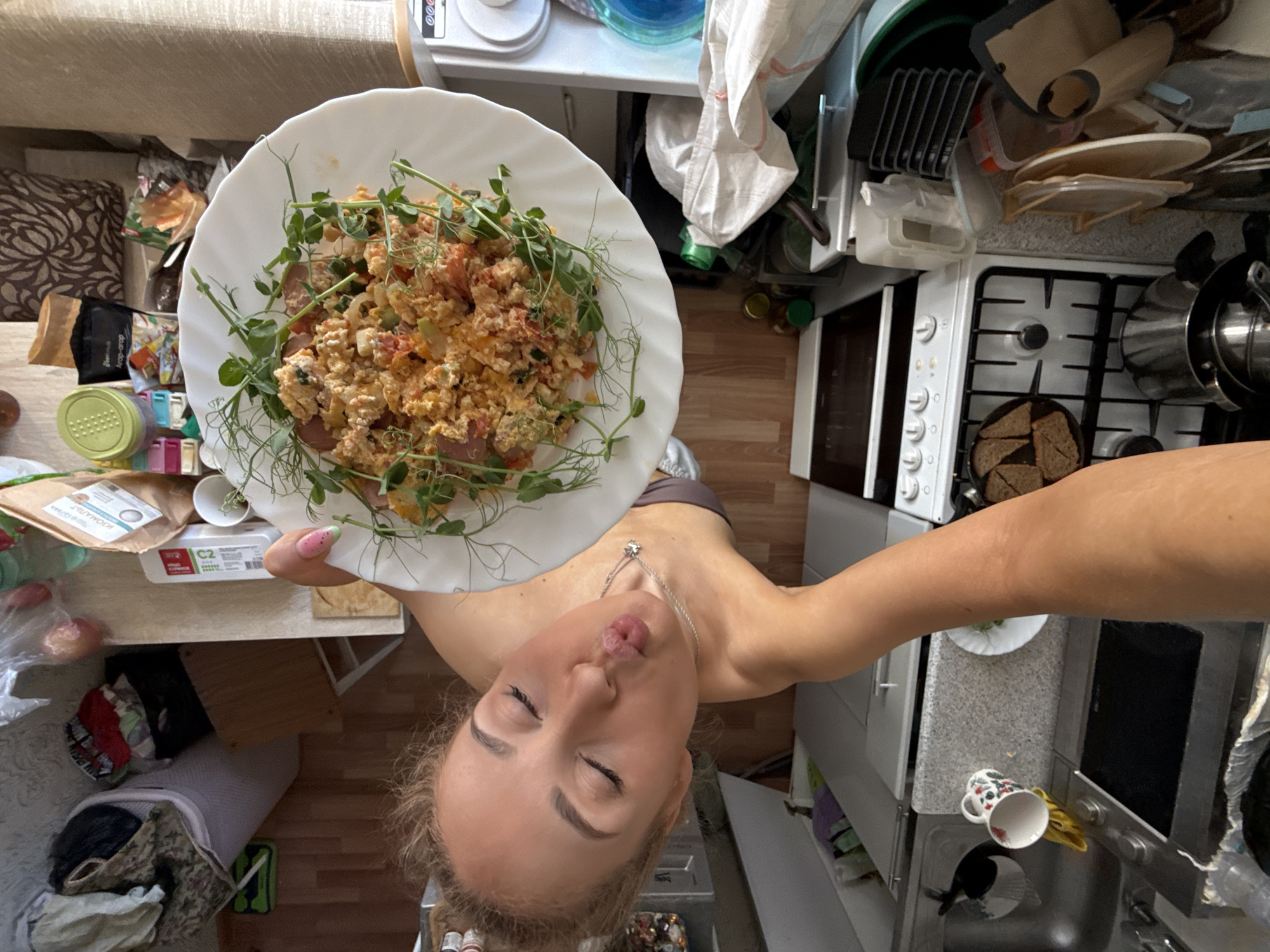 A woman is taking a selfie in a kitchen, holding a plate of food garnished with greens. The food appears to be scrambled eggs with tomatoes. The kitchen appears cluttered with various items on the counters and on the stove. Items include a kettle, dishes, a microwave, and other kitchen appliances. The city is not discernable in the image.