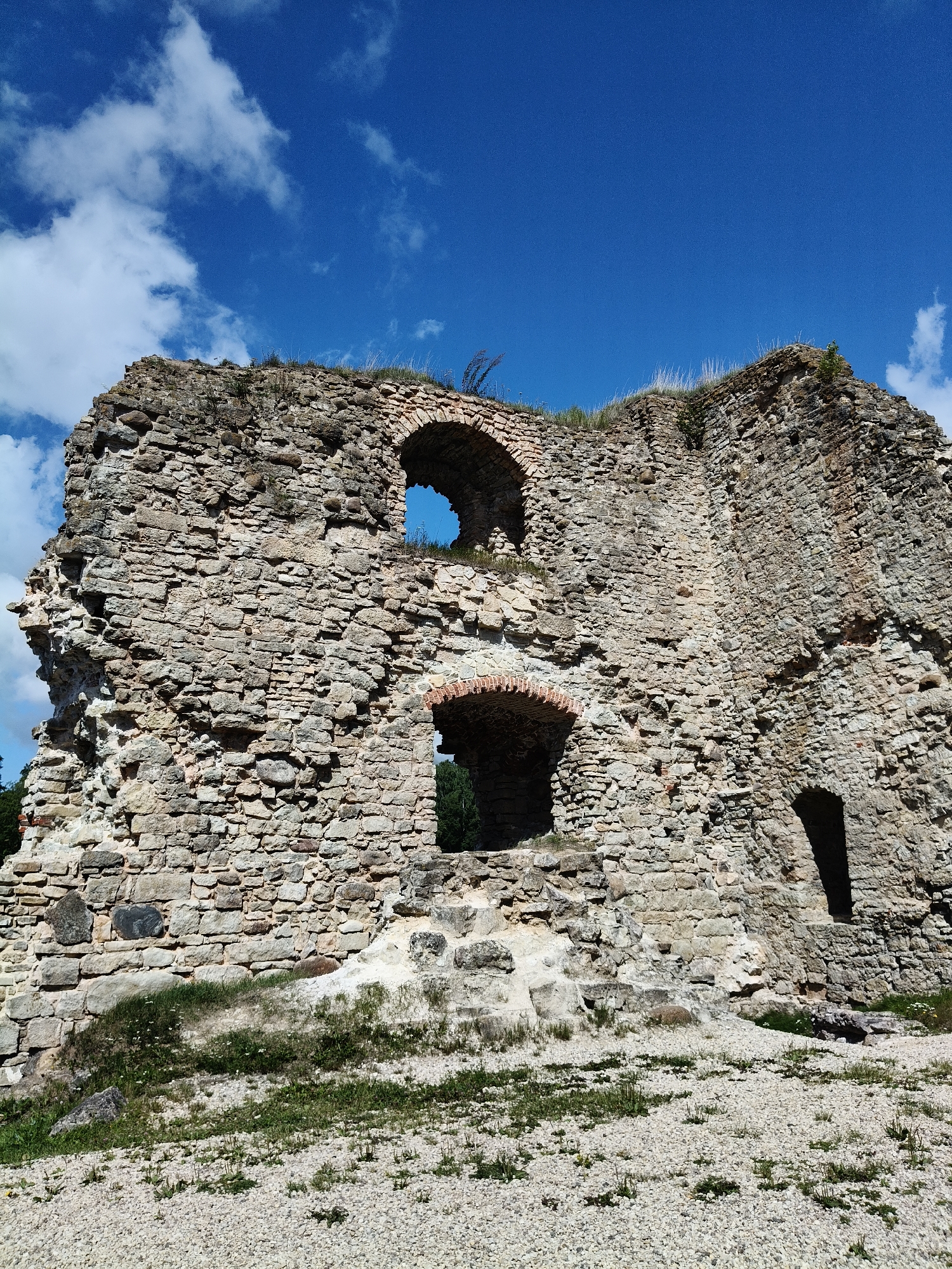 The image shows the ruins of a stone castle in Koknese, Latvia. The sky is blue with scattered white clouds. The castle walls are made of rough, uneven stones and show signs of age and decay. There are several arched openings in the walls, some of which are dark and lead to unknown interiors. The ground in front of the castle is covered with gravel and patches of grass. The overall scene suggests a historical site that is partially preserved and open to the elements.