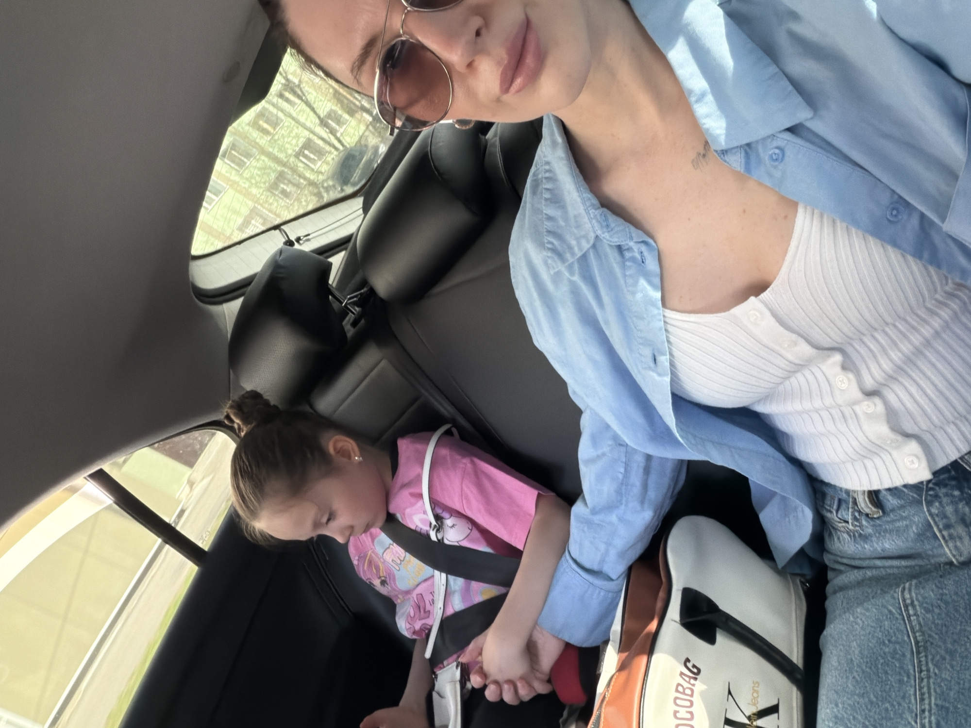 The photograph, taken inside a car, depicts an adult and a child. The adult, with sunglasses perched on their face, is positioned in the front passenger seat, oriented towards the camera. They are wearing a light blue shirt over a white top, and blue jeans are visible. A handbag is placed near the adult. The child, in the back seat, is wearing a pink shirt and is secured with a seatbelt. The background reveals glimpses of an urban environment. The photograph was taken in Perm, Russia.