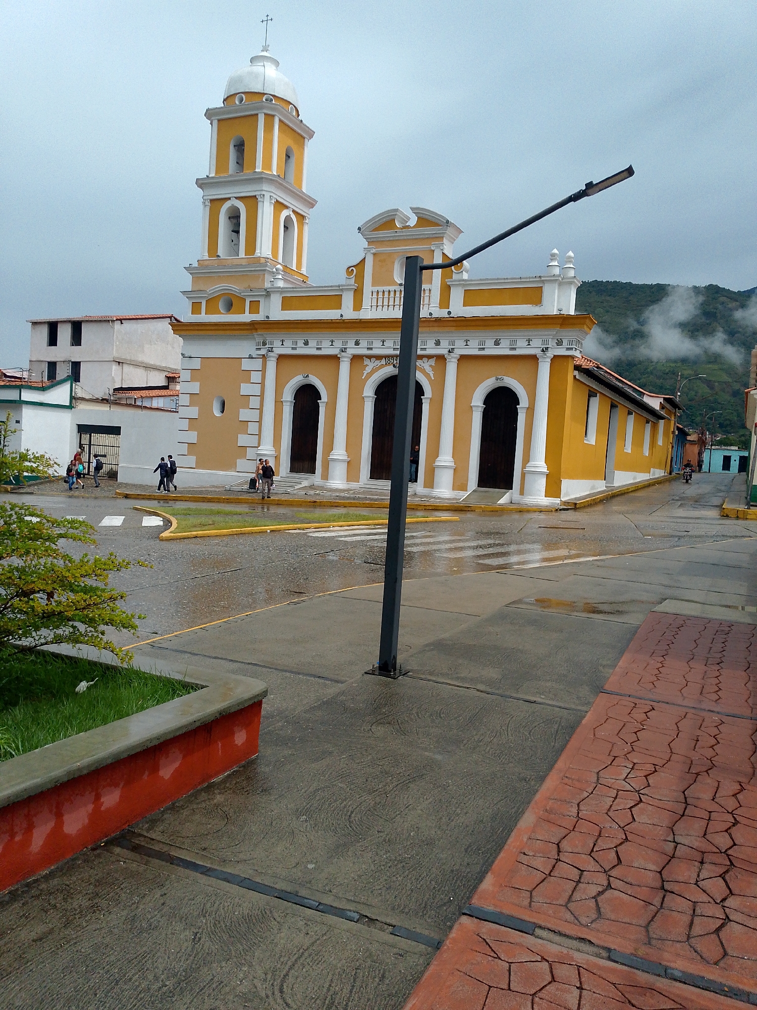 The image captures a street view in Mérida, Venezuela, focused on a prominent yellow and white church with a bell tower. The church's facade features arched doorways and classical columns. The surrounding streets are wet, suggesting a recent rain. Several people are walking across the street near a crosswalk. A street lamp stands in the foreground, partially obscuring the view. In the background, mountains are visible, partially obscured by clouds. A small patch of greenery is present on the left, bordered by a red wall. The sidewalk on the right is paved with patterned red bricks. The scene appears to be a town square or central location in Mérida.