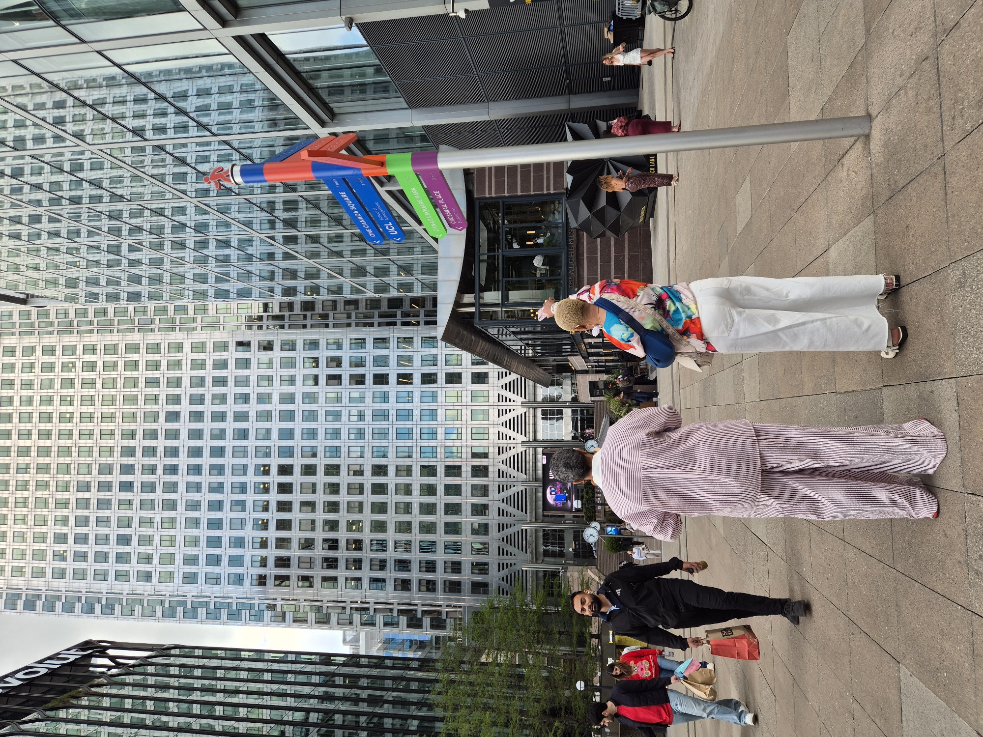 Here is a description of the image:
The image shows a busy urban scene in Canary Wharf, London, United Kingdom. In the center-left stands a tall, modern building with a grid-like facade of windows reflecting the sky. A colorful directional signpost is prominent, with arrows pointing towards "One Canada Square," "UCL," "Canada Square Park," and "Crossrail Place," indicating the landmarks nearby.
Pedestrians populate the scene, including two individuals in patterned outfits, one wearing a brightly colored floral print and white pants, and another wearing a striped suit. A man in a black tracksuit and face mask walks past, carrying a shopping bag. Other people are scattered in the background. There are also signs for “The Alchemist” and "CK Lane".