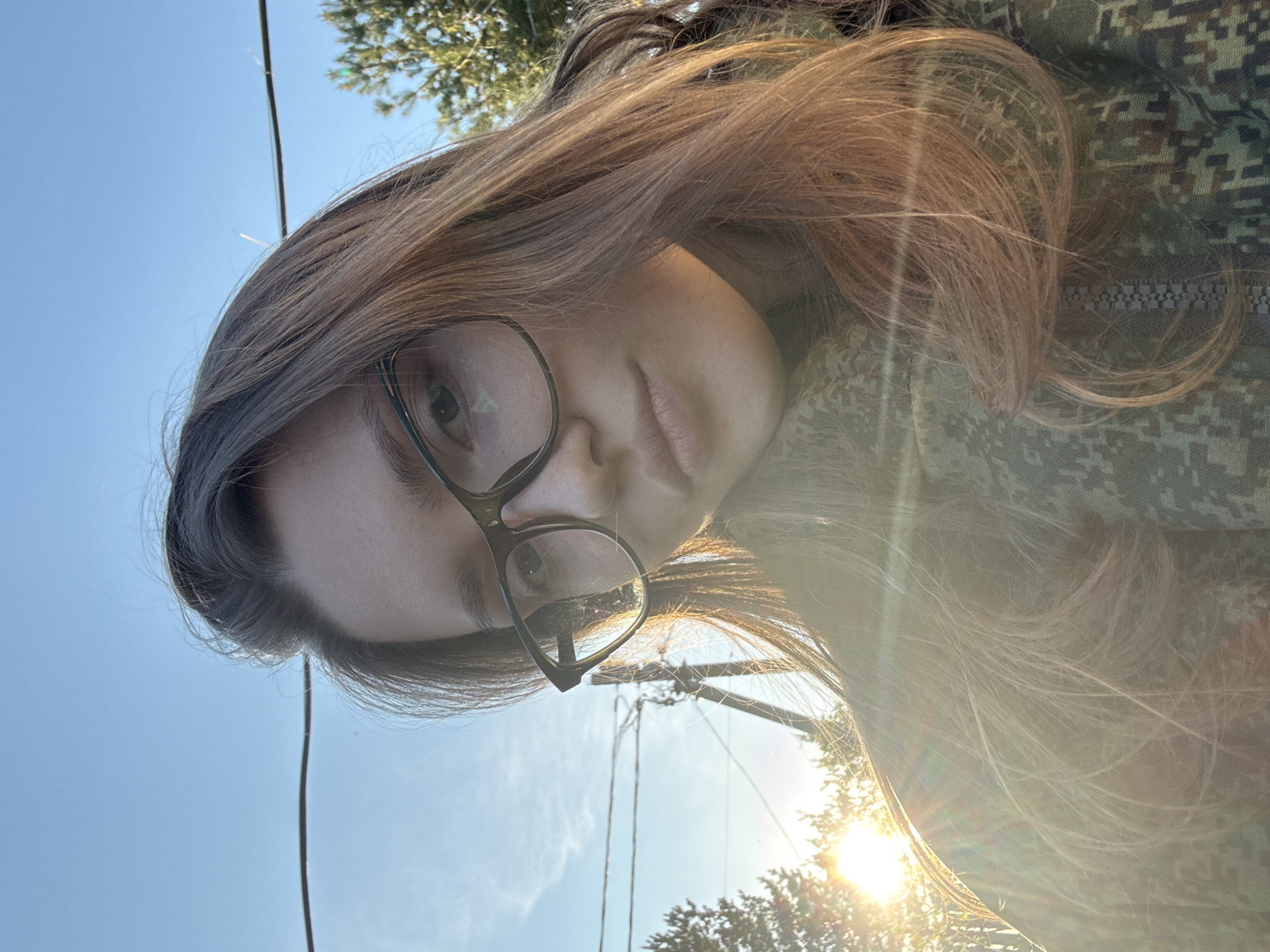A photograph shows a woman wearing eyeglasses, with her face partially obscured by her long, brown hair. She is wearing a camouflage-patterned garment. The sun is shining brightly, causing lens flare. In the background, there is a view of the sky, trees, and power lines. The image appears to be taken outdoors in Ленинградская, Russia.