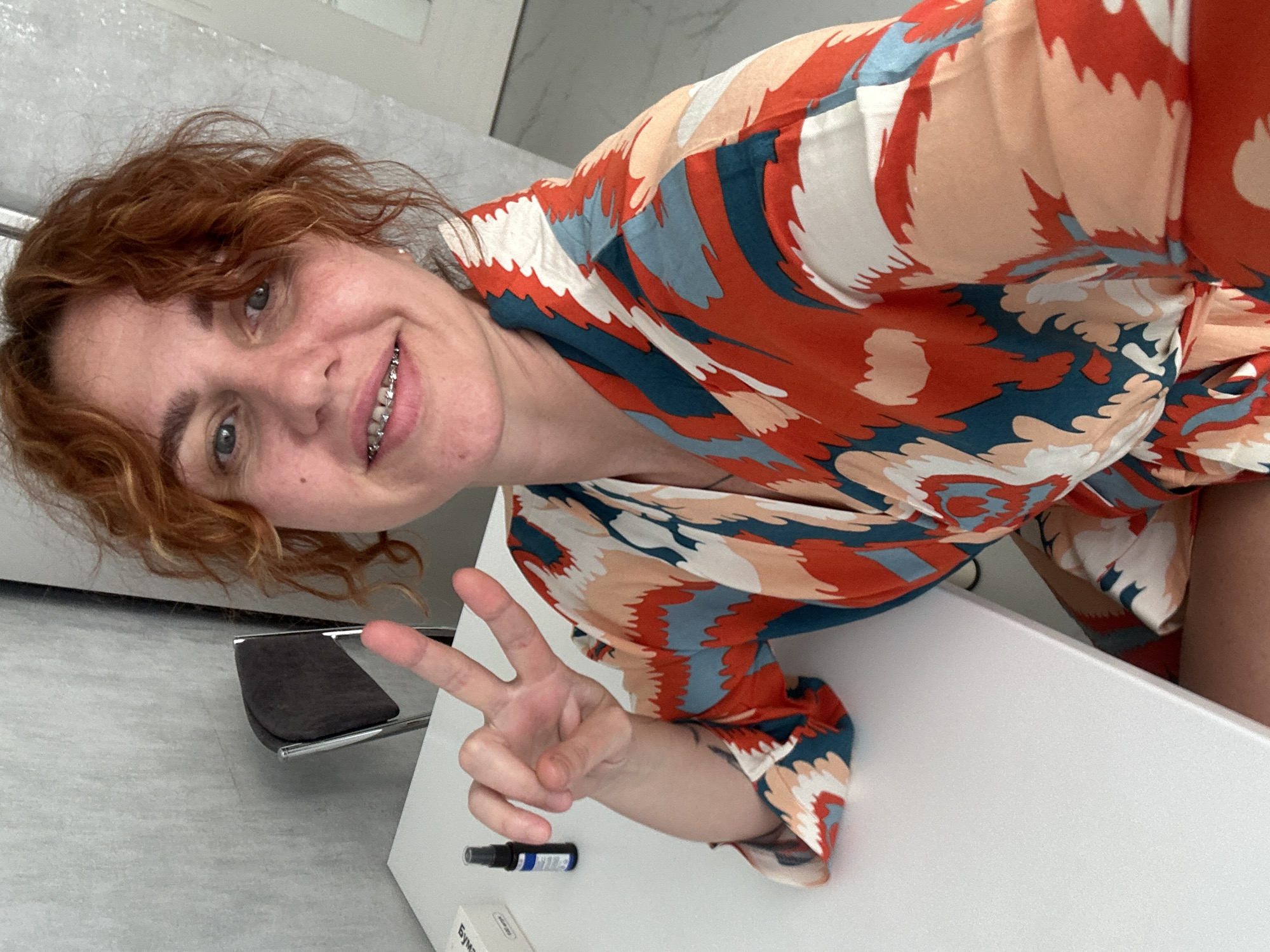 A woman with red curly hair and braces is making a peace sign. She is wearing a colorful orange, blue, and white patterned shirt. On the white surface next to her is a small black spray bottle and a partially visible box labeled "Бумага-30". The background appears to be a room with a grey floor. The location is Sochi, Russia.