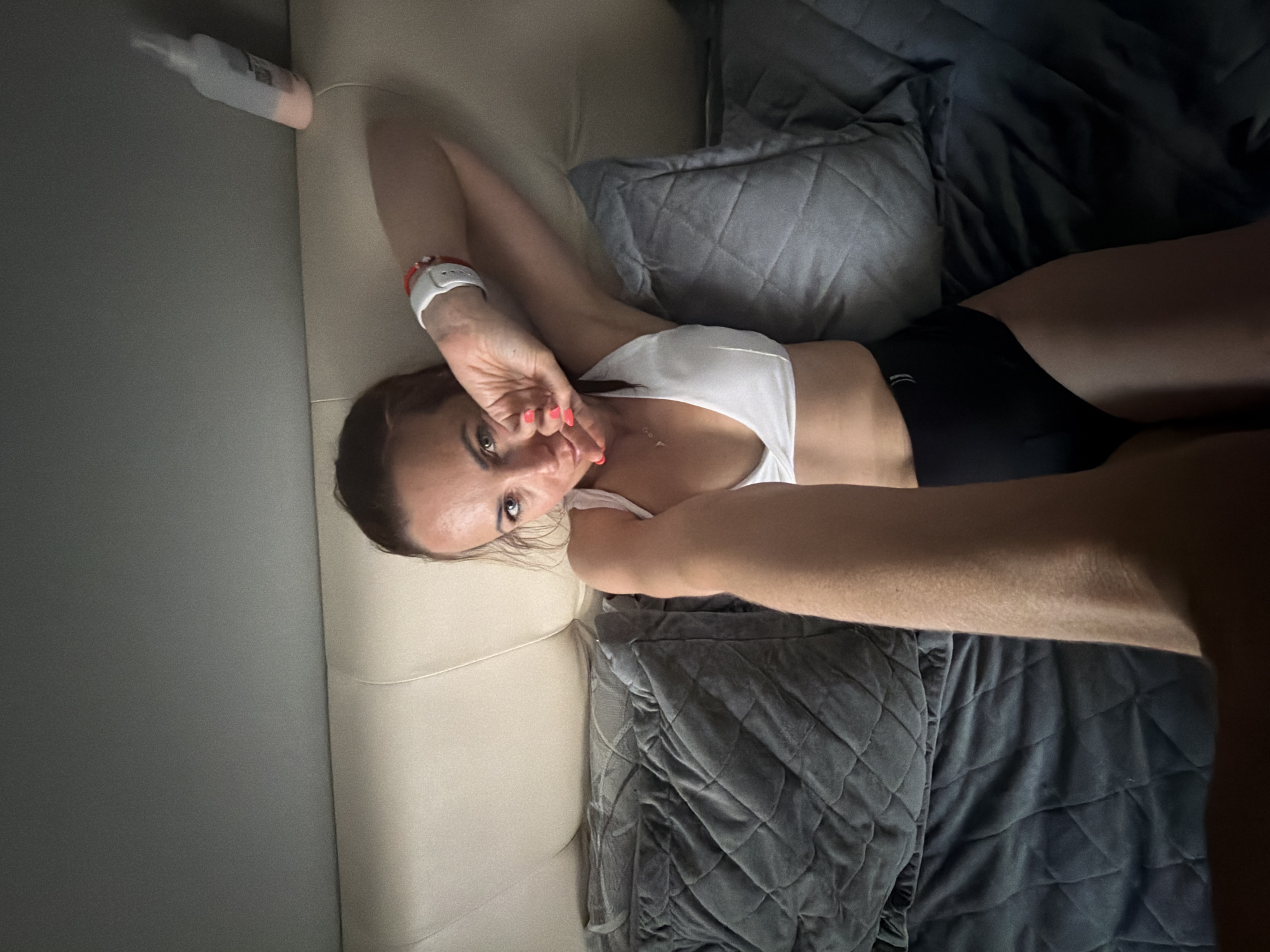 A woman lies on a bed with a cream-colored headboard and gray blankets. She is wearing a white sports bra and black bottoms. Her left arm is extended towards the camera, and her right arm is bent at the elbow, with her hand near her face. A white bottle with a pink cap is visible on the headboard in the background. The scene appears to be indoors, in a bedroom.