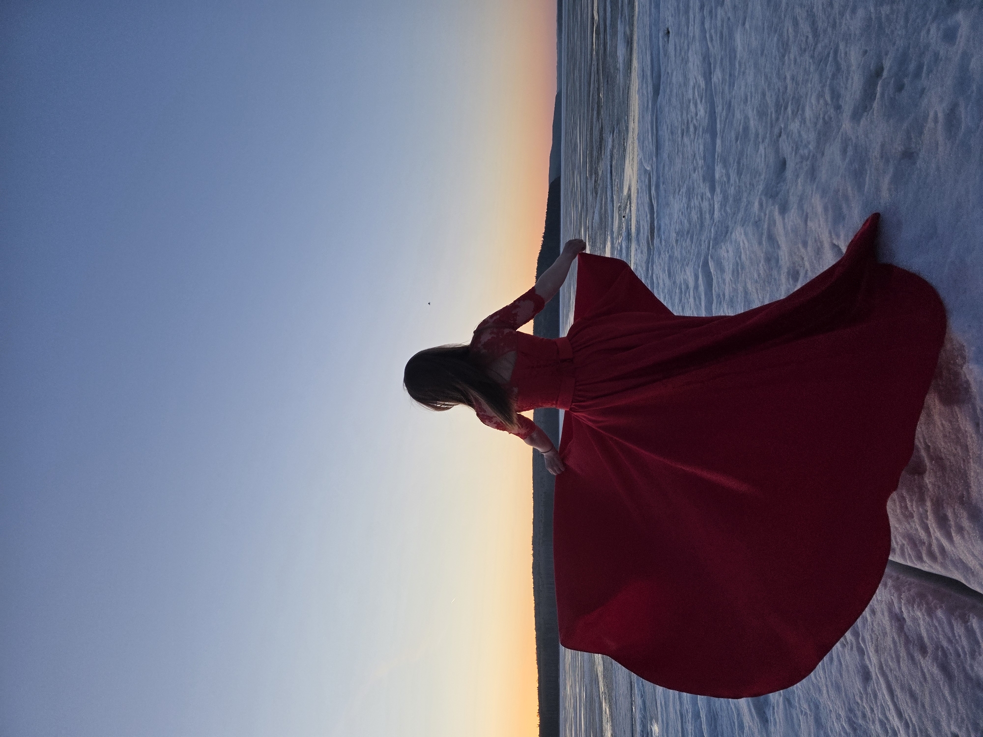 A person wearing a red dress is seen with outstretched arms in a landscape under a dusky sky. The location appears to be Chaikovsky, Russia. The ground is covered in snow and what seems to be a frozen river or lake. The sky transitions from blue to orange as the sun sets.