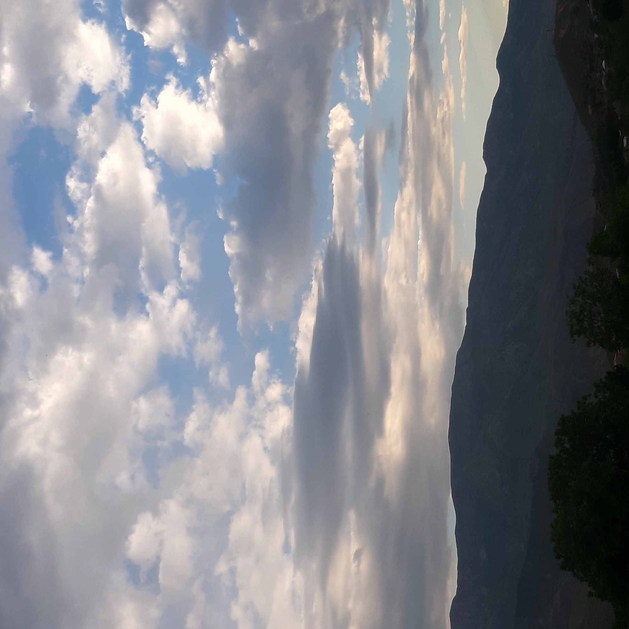 The image captures a serene mountainous landscape under a dynamic sky, likely during late afternoon or early evening. The majority of the frame is filled with a vast expanse of sky, featuring a mix of cumulus and stratocumulus clouds. These clouds display varying tones, from bright white where illuminated by sunlight to darker shades of grey, with patches of clear blue sky visible between them.
On the right side of the composition, a large mountain range forms a dark silhouette against the lighter horizon, its prominent, undulating ridgeline creating a striking profile. The mountain's dark appearance suggests it is cast in shadow, possibly from a low-lying sun. In the bottom right corner, dark green foliage, indicative of trees, adds a natural foreground element, partially obscuring the base of the mountain.
No people or specific activities are depicted, focusing solely on the natural environment. The scene conveys a sense of grandeur and peacefulness. Given the contextual information, this view is characteristic of the mountainous region around Yeghegnadzor, Armenia. There is no visible text in the media file.