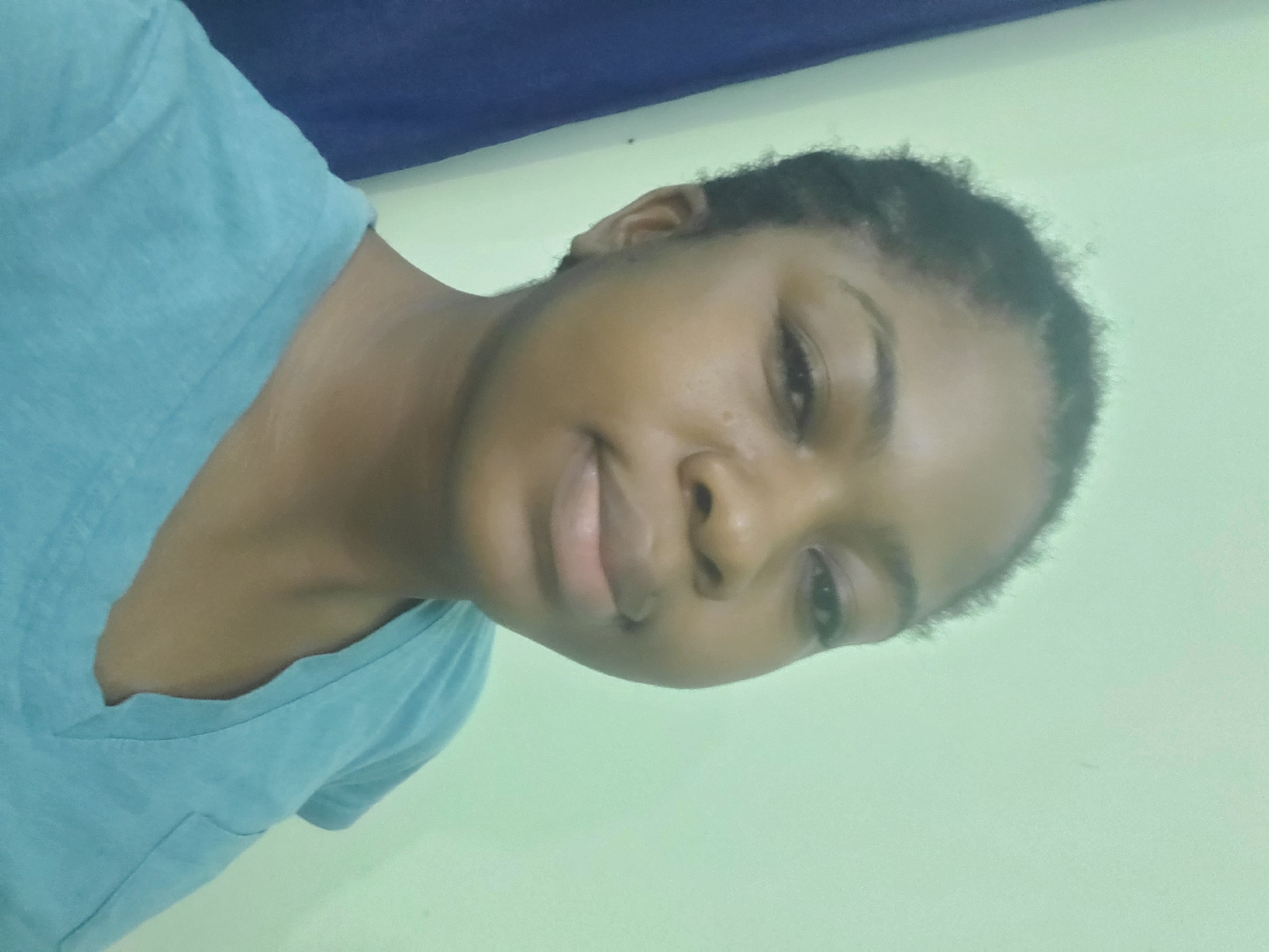 A young Black woman is pictured from the shoulders up, facing the camera. She has dark, short, braided hair pulled back from her face. Her skin tone is medium brown. She is wearing a light blue V-neck t-shirt. The background is a plain, light green wall. To the left of the woman, a dark blue curtain or piece of fabric is visible. The lighting is even, and the image appears to be taken indoors, likely in a residential setting in Barden Yana, Nigeria.