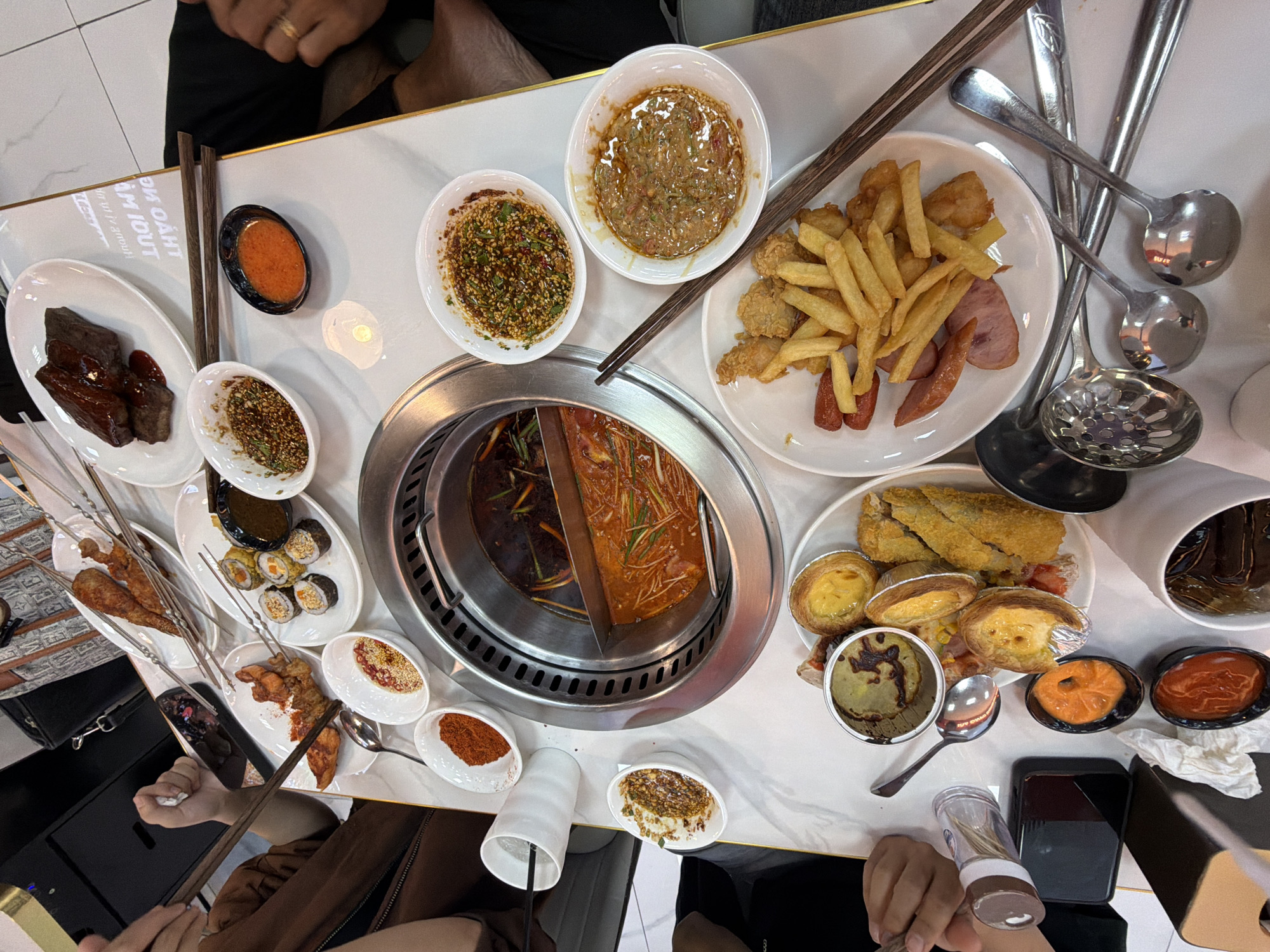 The image depicts a dining table laden with various dishes, sauces, and utensils, typical of a hot pot or communal dining experience. The central feature is a divided hot pot with dark and reddish broths. Plates surrounding the hot pot contain fried foods, skewers, sushi, and other unidentified dishes. Several small bowls hold sauces, spices, and condiments. Utensils such as chopsticks, spoons, and ladles are scattered around. People are partially visible around the table, suggesting a group dining setting. Based on the style of food and the possible presence of Vietnamese script on a plate in the background, the location is likely in Đà Nẵng, Vietnam.