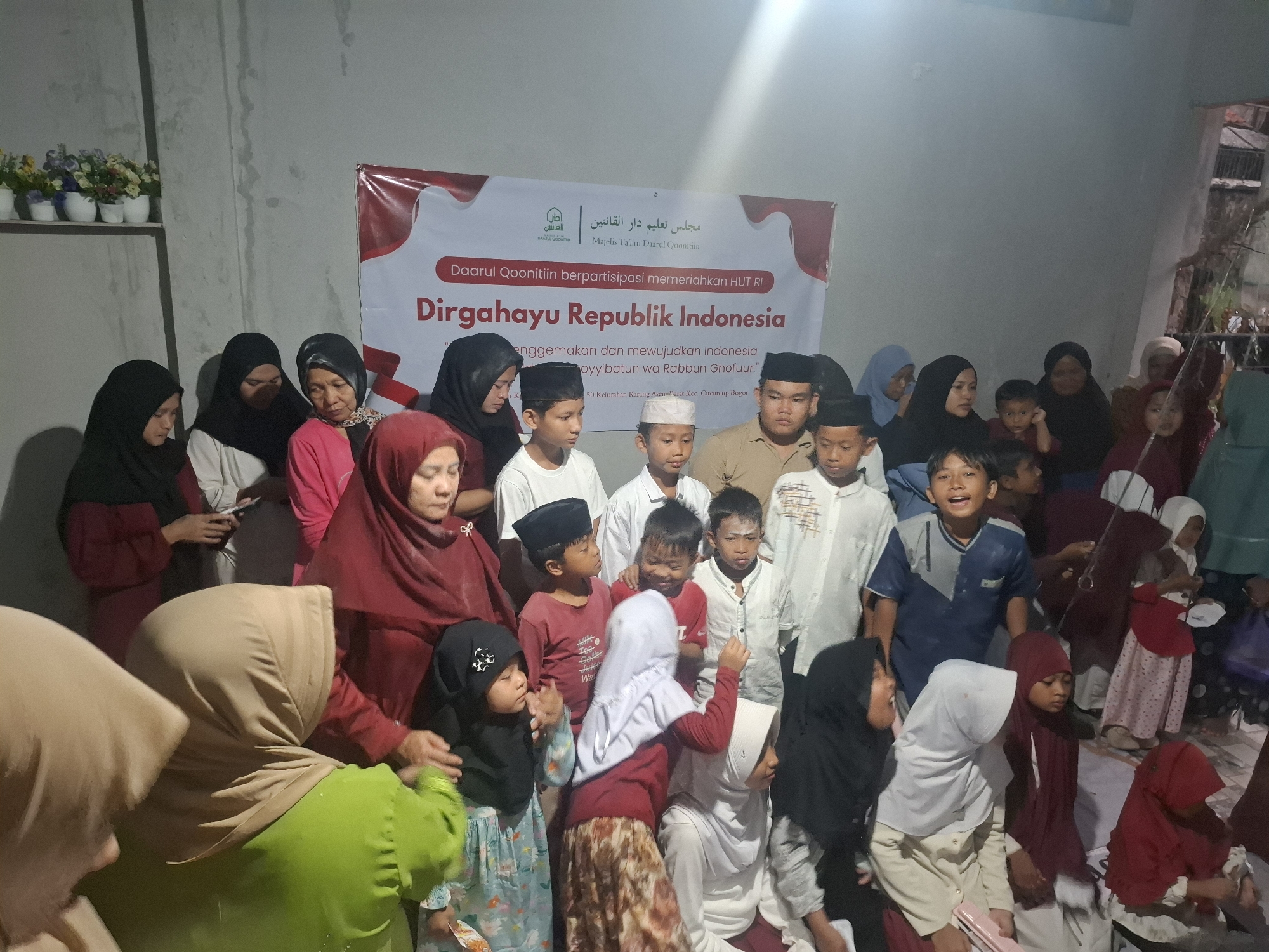 In an indoor gathering, a group of people, primarily women and children, are present. A banner in the background, featuring Arabic calligraphy and text in Indonesian, reads "Darul Qoonitiin berpartisipasi memeriahkan HUT RI Dirgahayu Republik Indonesia" and mentions "Majelis Ta'lim Darul Qoonitiin" and an address in Babakan Madang, Bogor, Indonesia. Several individuals are wearing Islamic attire, including hijabs and skullcaps. Some of the children appear to be interacting with each other, while others are looking towards the camera or away. The environment seems to be a simple interior, possibly a hall or a room, with a plain wall and some decorations on a shelf in the upper left corner. The lighting is somewhat dim, with a bright light source from the upper right casting shadows.