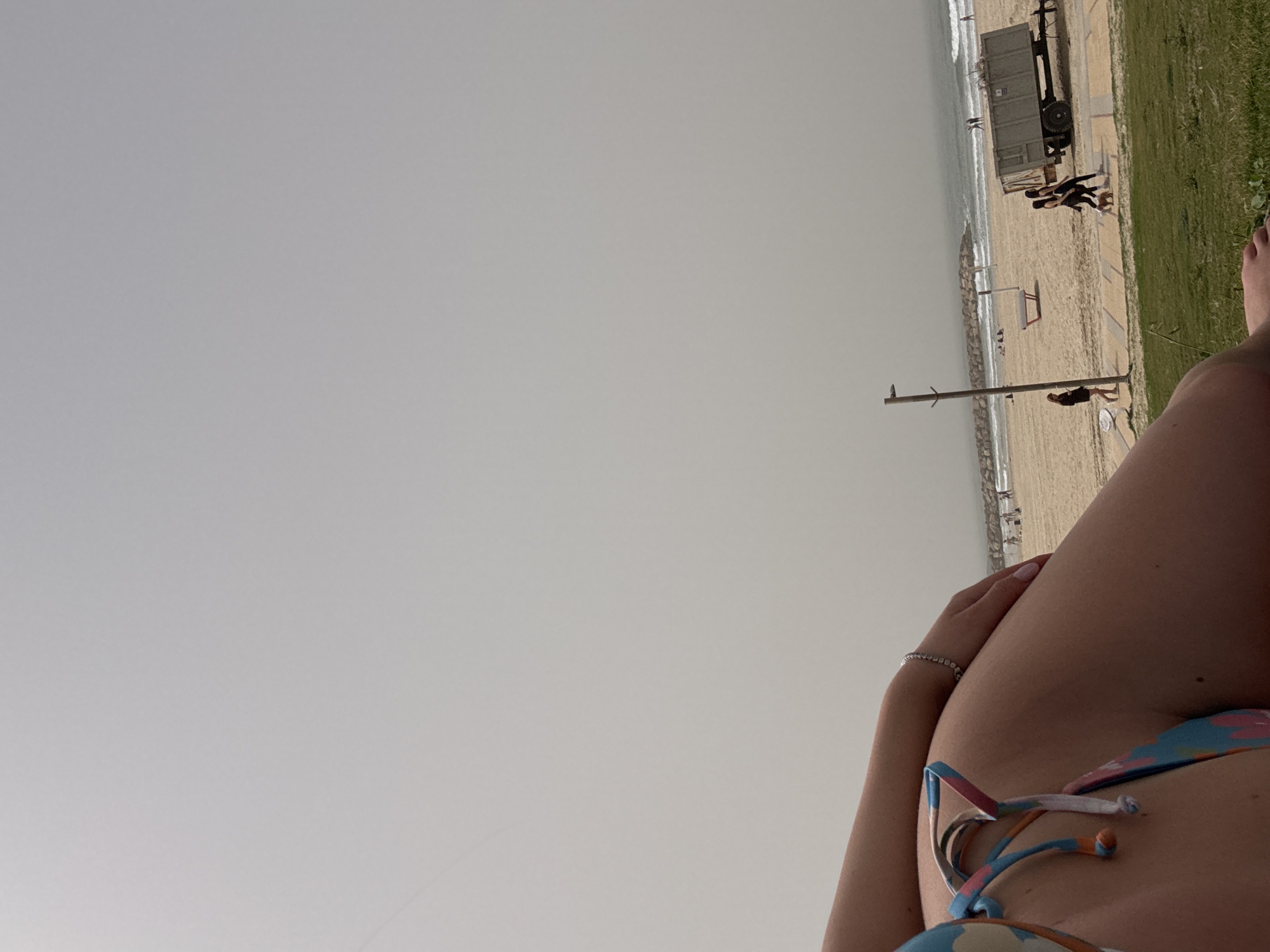 View of Herzliya beach, Israel, from a prone person's perspective. The sky is overcast. The beach is visible with the ocean, sand, people, and a truck parked nearby. The person is wearing a bikini and a bracelet.