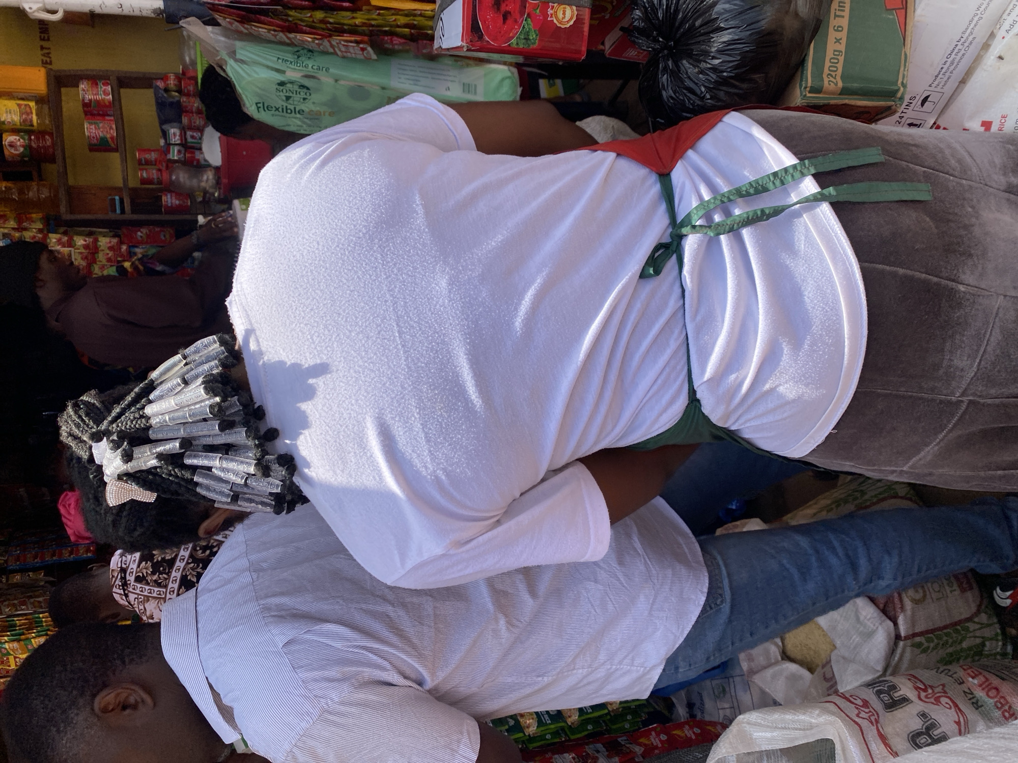 This is a medium shot of people in a marketplace in Jos, Nigeria. In the foreground, two individuals are visible from the back. The person on the left wears a white striped shirt and blue jeans. The person on the right wears a white t-shirt with a green apron tied around their waist and a long brown skirt. Their hair is styled in braids with clear plastic accessories. In the background, a person is seen behind a stall stocked with canned goods and other items. Several bags of rice are stacked near the foreground subjects. The overall environment suggests a bustling marketplace setting.