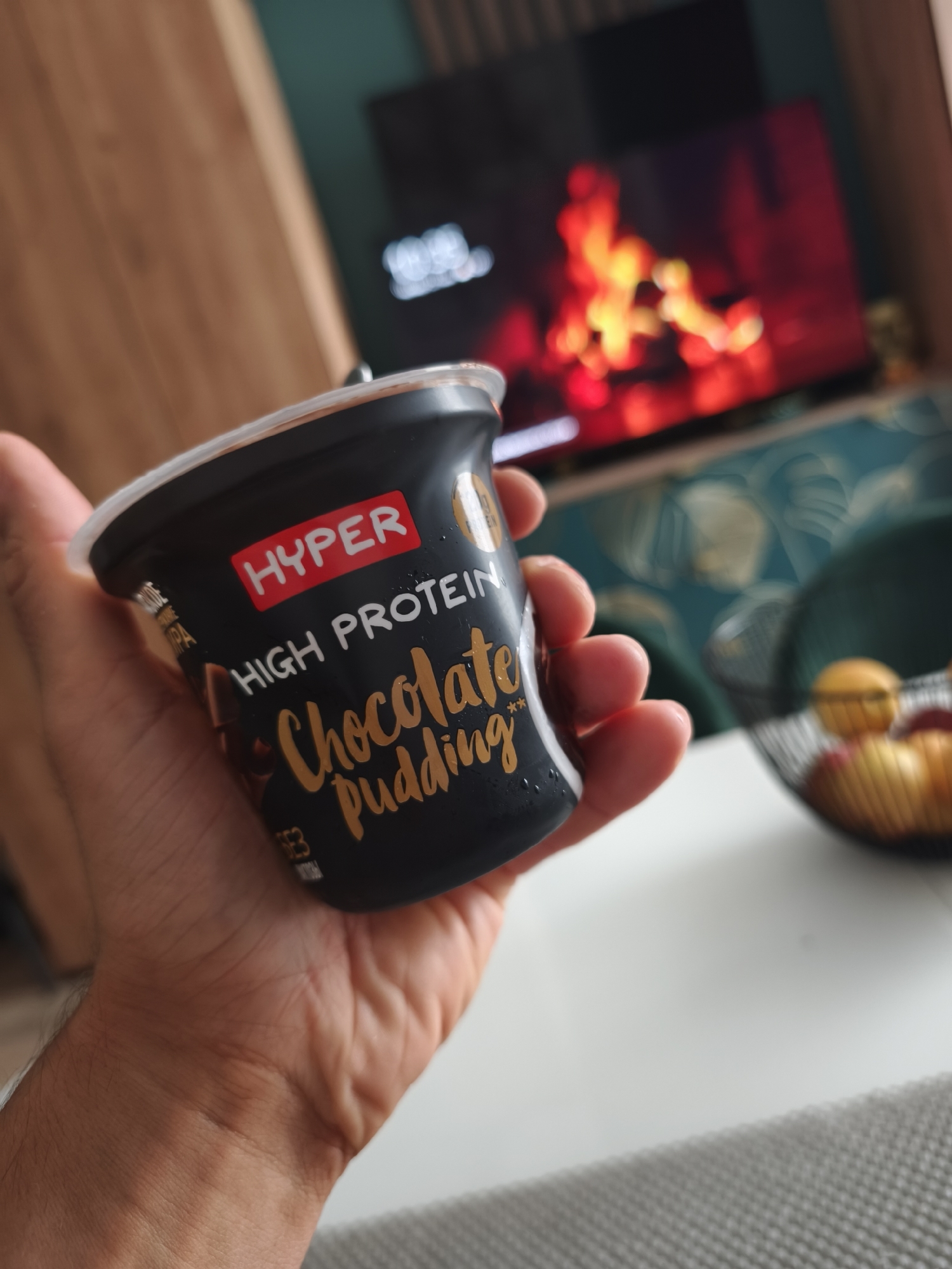 A hand holds a container of "Hyper High Protein Chocolate Pudding" indoors. A television displaying a fireplace scene is visible in the background. A fruit basket sits on a white table. The environment suggests a living room setting. The location is not discernible from the image and cannot be accurately identified as Moscow, Russia.