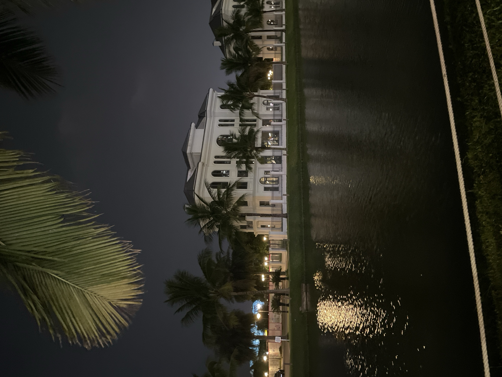In Hà Nội, Vietnam, at night, a large, white, multi-story building with a classical architectural style stands prominently on the far bank of a dark body of water. The building features numerous windows, arched doorways, and decorative columns, and is illuminated by artificial lights. Palm trees with their fronds silhouetted against the dark sky are visible in front of and around the building, as well as in the foreground. The water reflects the lights from the building and the surrounding area, creating shimmering patterns on its surface. A white rope barrier with grass behind it runs along the edge of the water in the foreground. The overall scene is serene and somewhat opulent, typical of upscale residential or resort areas in Hà Nội.
