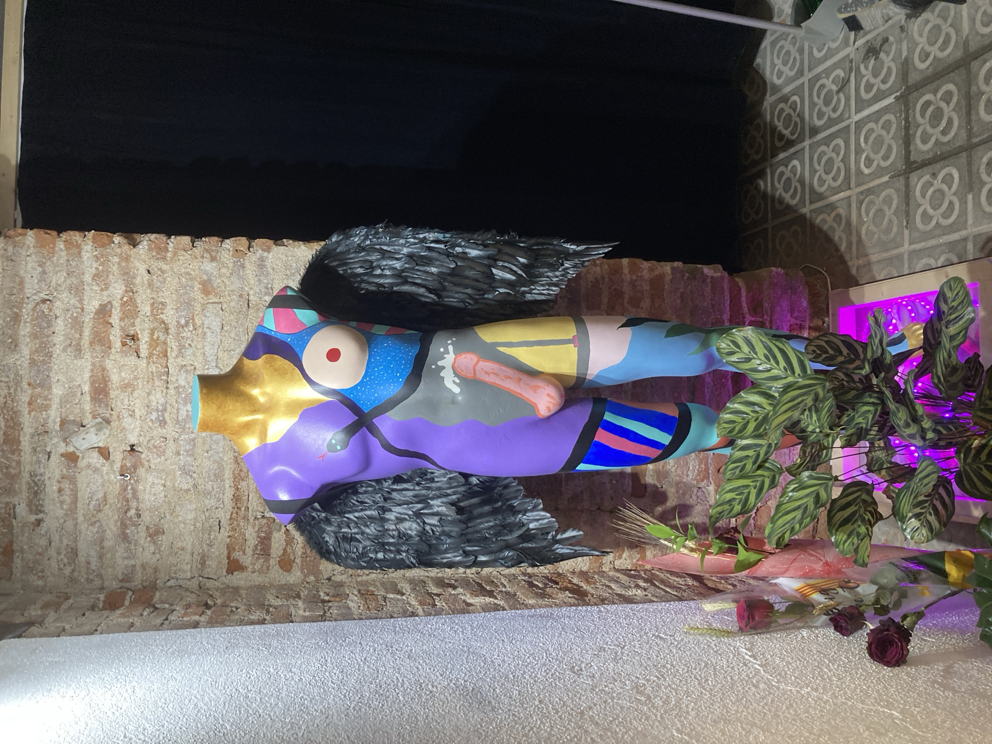 An artistic mannequin or statue, painted in vibrant, abstract designs, stands prominently in a dimly lit setting. The figure, resembling a female torso and legs, features a golden neck, one red circular breast, and a black nipple on the other. A prominent light pink phallic symbol is painted on its abdomen. Black, feathered wings are attached to its back, and a black snake design coils on a purple section of its body. The figure is adorned with various patterns and colors including blue, purple, yellow, pink, red, and black stripes.
The scene appears to be an interior or covered outdoor space, possibly at night, with a dark area visible through an opening in the upper left, suggesting a window or doorway. A rustic brick wall forms the backdrop on the left and central parts of the image. To the right, a distinctive tiled wall featuring a repeating floral or quatrefoil pattern, characteristic of Barcelona's "Panot de flor" tiles, is visible.
On the right side of the statue, a lush green plant with striped leaves is illuminated by a striking purple light emanating from behind it. On the ground in front of the plant, several cut flowers and decorative items are arranged, including a few red roses (some appearing dried), stalks of wheat, and an item wrapped in clear plastic. The floor is a light, textured surface. No specific activity is taking place, as it's a static display. There is no clearly legible text visible in the image.