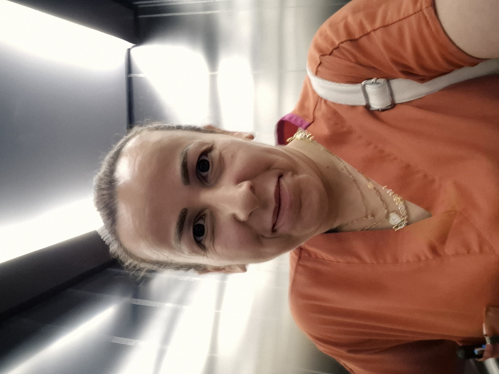 A woman with dark hair pulled back, wearing an orange scrub top and a white strap across her shoulder, smiles as she takes a selfie in an elevator. She is wearing two necklaces, one with a heart pendant. The elevator has reflective walls and bright overhead lighting.