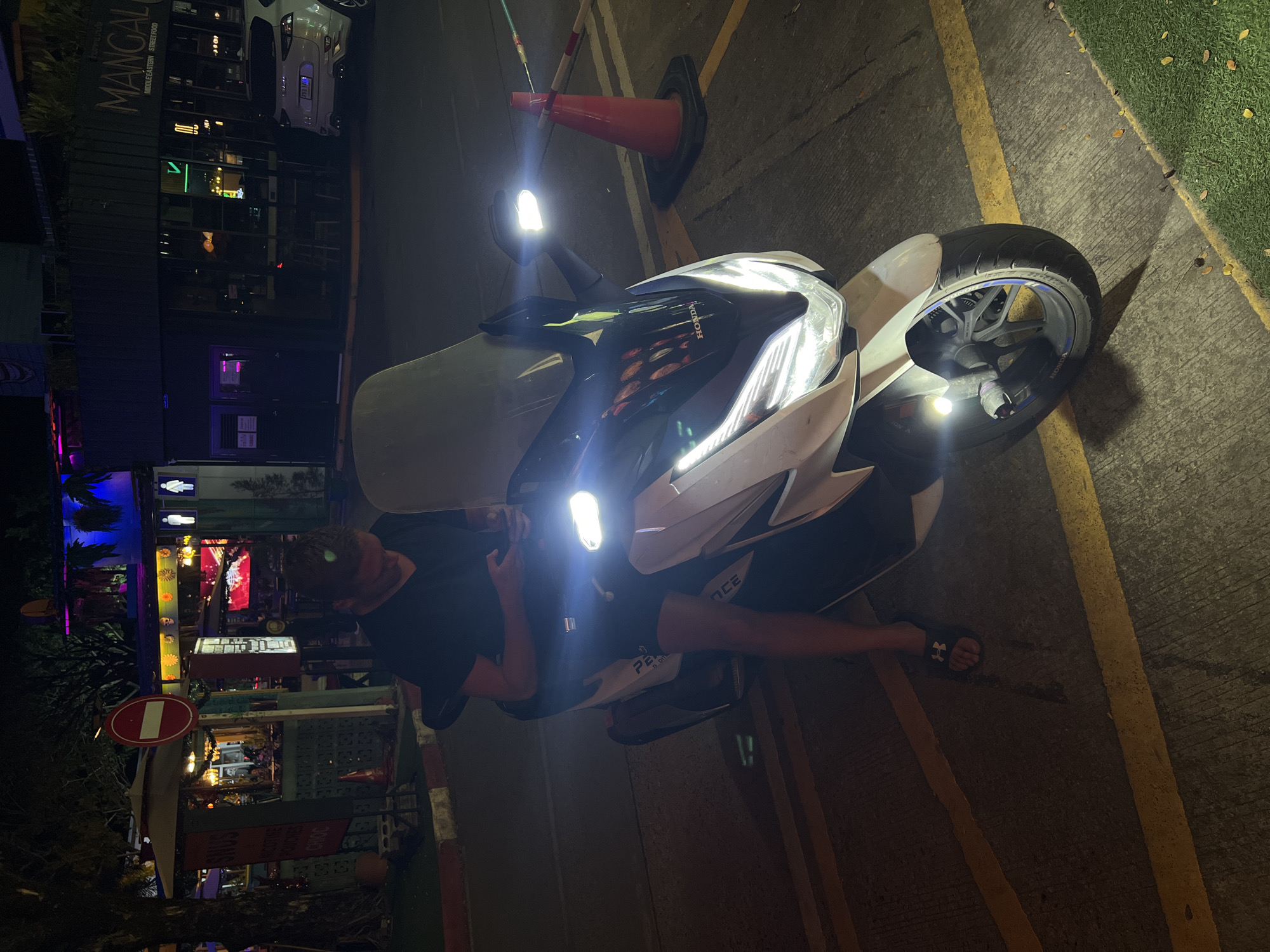 A high-angle, slightly rotated view captures a well-lit scene. The primary subject is a parked white and black Honda scooter, facing the upper right of the frame. Its headlights are on, casting bright light. A person, partially visible, stands near the scooter, wearing a black t-shirt and shorts, and is engaged with the vehicle. An orange traffic cone sits behind the scooter's front. To the left, a street with colorful signage and building facades suggests a commercial area, possibly at night. The street exhibits various signs and decorative elements. "MANGAL..." is legible on one of the buildings. To the right, a section of pavement transitions to a grassy area. The location appears to be an urban setting, specifically a parking area adjacent to a vibrant street with commercial establishments. The image is slightly overexposed due to the bright headlights.