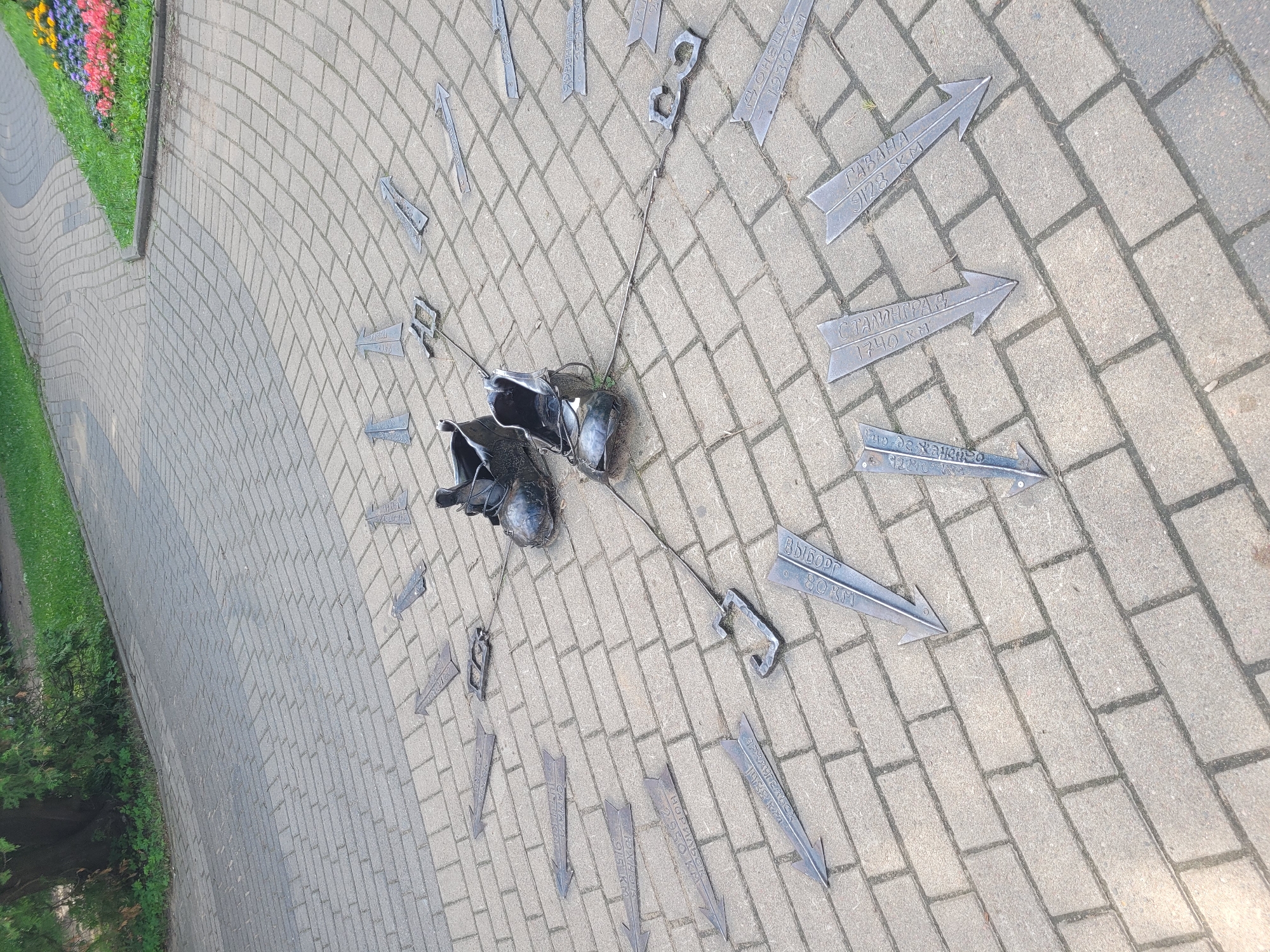 The image shows a pedestrian path paved with rectangular gray bricks, forming a circular pattern. Two weathered, dark-colored boots are placed in the center of the circle. Several metallic arrows, each pointing outwards from the center, are embedded in the brickwork at intervals, resembling the markings of a sundial or a directional marker. Some of these arrows are inscribed with text, likely place names and distances. For example, one arrow clearly reads "ГАВАHA 9128 KM" and another "СТАЛИНГРАД 1740 KM". This art installation is located in the city of Zelenogorsk, Russia, likely in a park or public square. A paved walkway curves through the background, bordered by a grassy area and a flower bed with colorful flowers. A tree with dense green foliage stands to the left of the image. The scene is outdoors, and the lighting suggests it is daytime.