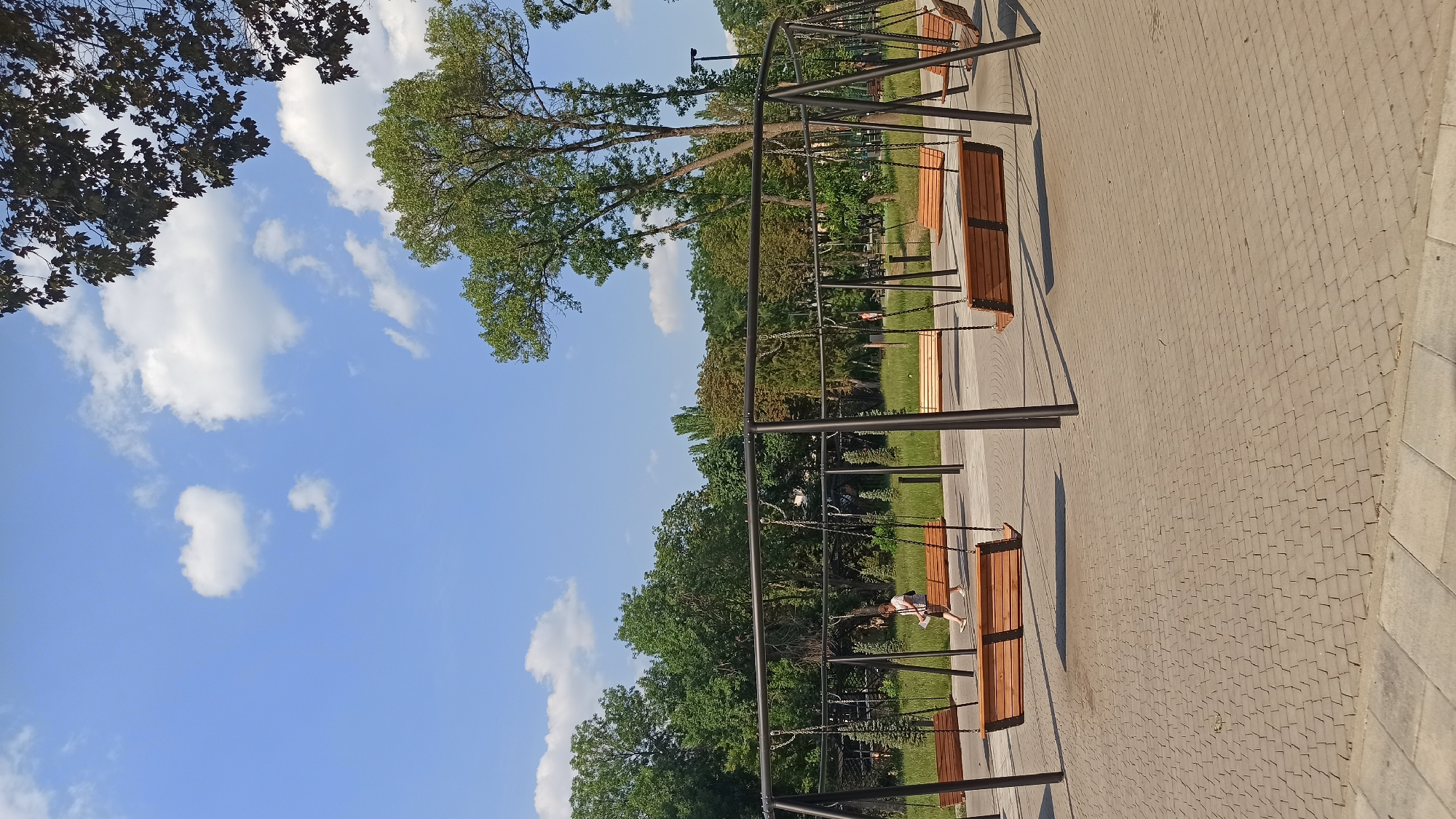 In an outdoor park, a person walks across a paved area with a series of modern wooden swings. The swings are suspended from dark metal frames that form an arc. The ground is covered in light-colored cobblestones laid in a circular pattern. In the background, lush green trees and a bright blue sky with scattered white clouds create a natural setting. The person, wearing light-colored clothing, is walking from left to right and holding a piece of paper. The scene suggests a peaceful, recreational area within a park.