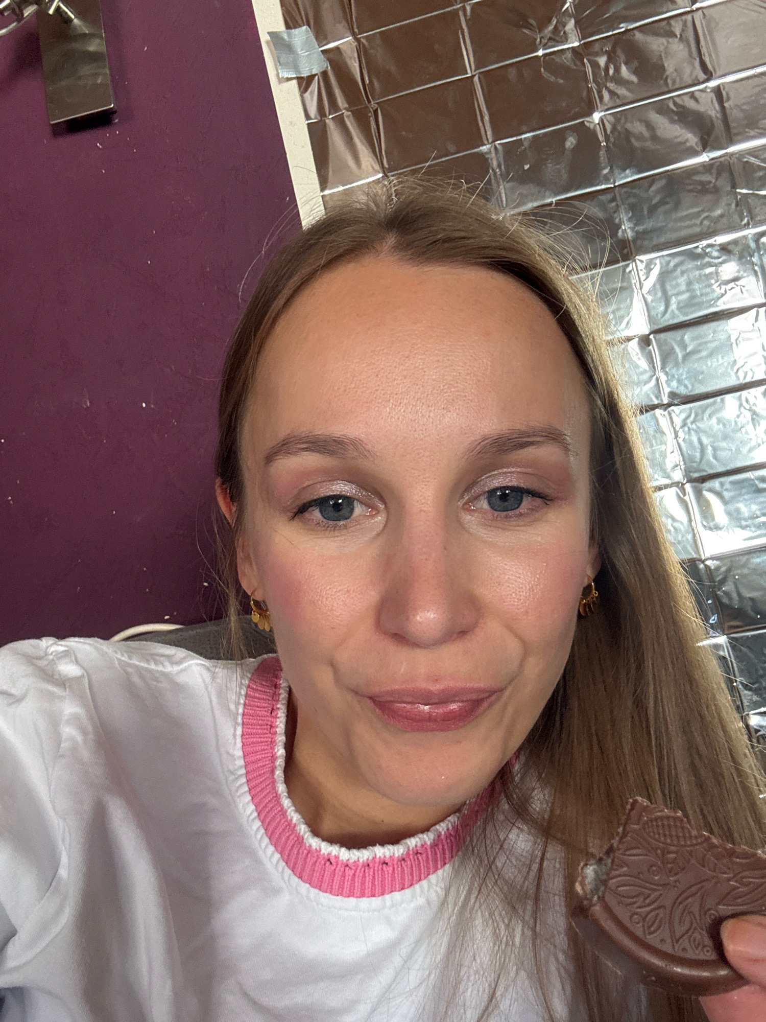 The image shows a close-up of a woman's face. She has long blonde hair, blue eyes, and is wearing makeup. She is smiling slightly and holding a piece of chocolate in her right hand. She is wearing a white shirt with a pink and white collar. The background is a wall with a metallic, tiled pattern and a purple section with some objects on it. There are no discernible city locations or specific details about Überherrn, Germany.