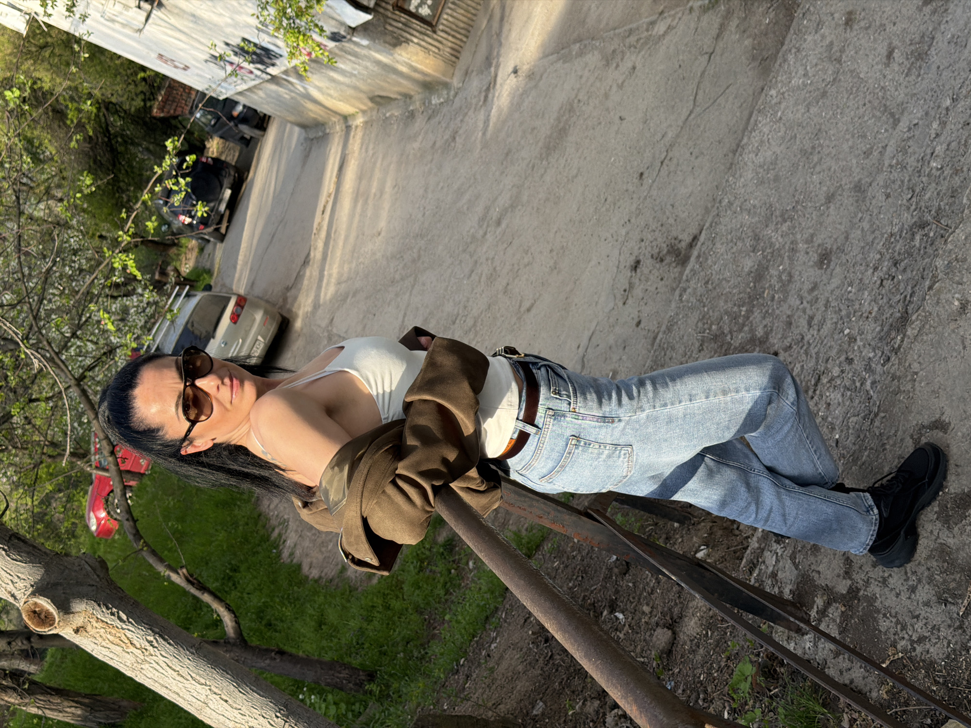A full-body portrait depicts a woman posing outdoors, leaning against a dark metal railing. She stands facing slightly right, looking towards the camera with a relaxed expression. She has long, dark hair pulled back and wears brown-tinted sunglasses. Her attire includes a white sleeveless top, light blue denim jeans, and black chunky-soled sneakers. A brown jacket is casually draped over her shoulders and arms, with the sleeves bunched up.
The scene is illuminated by strong, warm sunlight, casting distinct shadows and suggesting it is either late afternoon or early morning on a clear day. Behind the woman and the railing, the environment consists of dirt and green grassy patches, along with several tree trunks and branches. Some branches bear fresh green leaves and white blossoms. Further in the background, a concrete or dirt path features parked vehicles: a silver car on the right and a darker SUV deeper in the frame. A portion of a red vehicle is visible among the trees on the left. A light-colored, textured concrete building forms the upper background. The setting suggests an outdoor location in a residential or mixed-use area, consistent with Pleven, Bulgaria. There is no visible text in the image.
