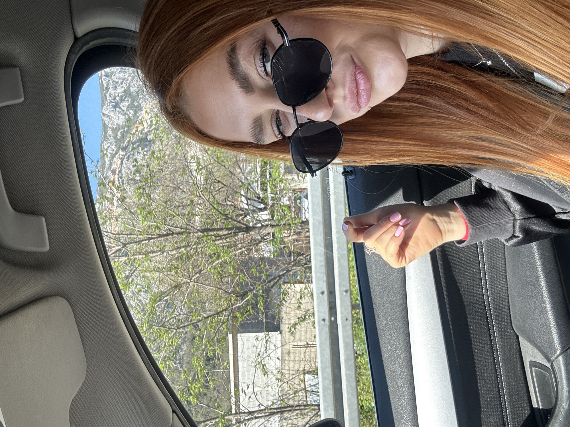 A woman with long auburn hair and black sunglasses is in a car. The landscape visible through the window includes mountains, trees, and buildings. The buildings appear to be in the urban area of Zgrade, Montenegro.