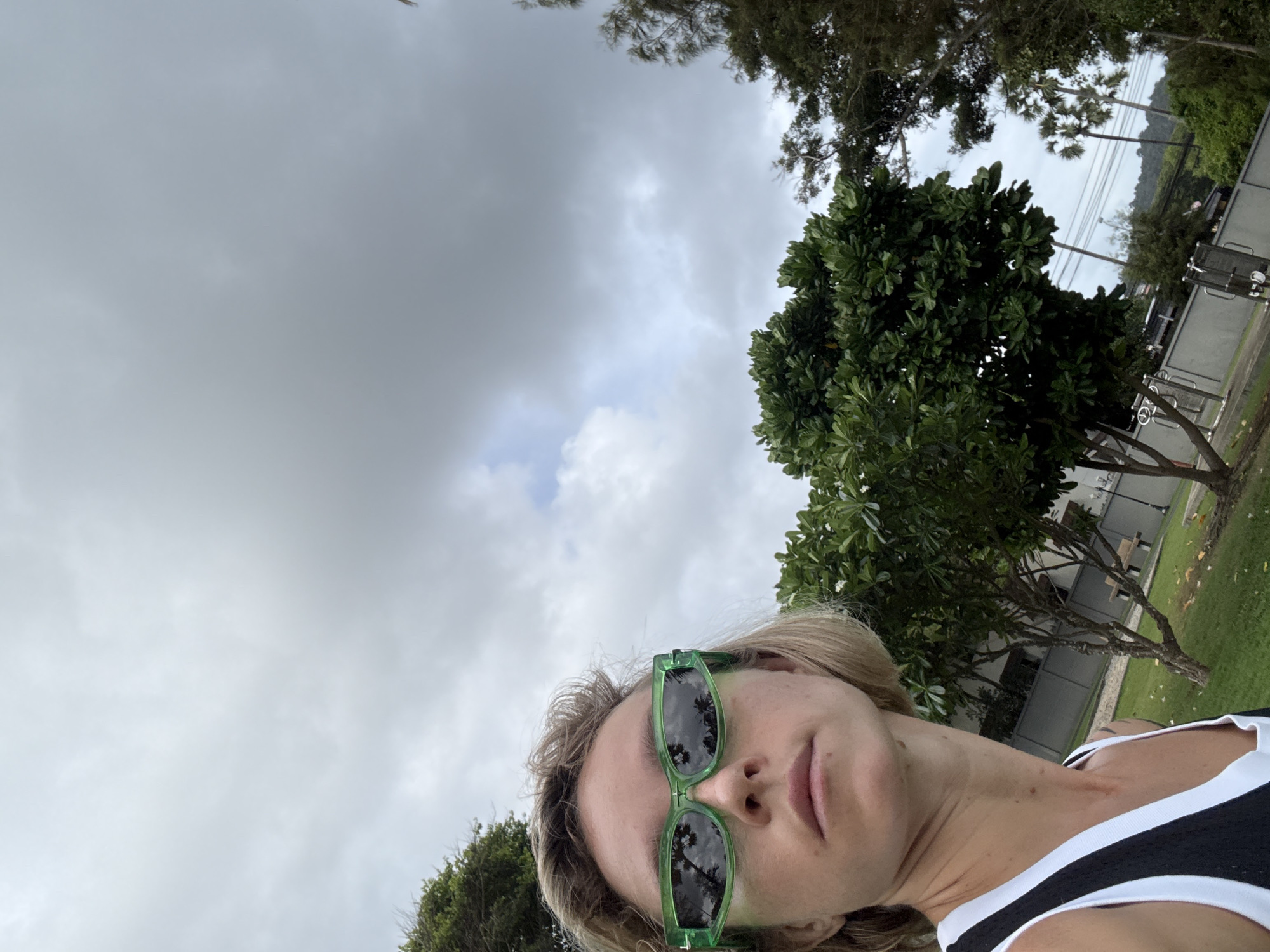 A close-up, low-angle shot shows a woman wearing large, bright green sunglasses and a black and white tank top. She has short, blonde hair and fair skin. The sunglasses reflect palm trees and a cloudy sky. The background features lush green trees, some with visible trunks, and a grey, overcast sky. In the distance, a concrete wall, benches, and what appear to be outdoor exercise equipment are visible. The overall setting suggests a park or outdoor recreational area.