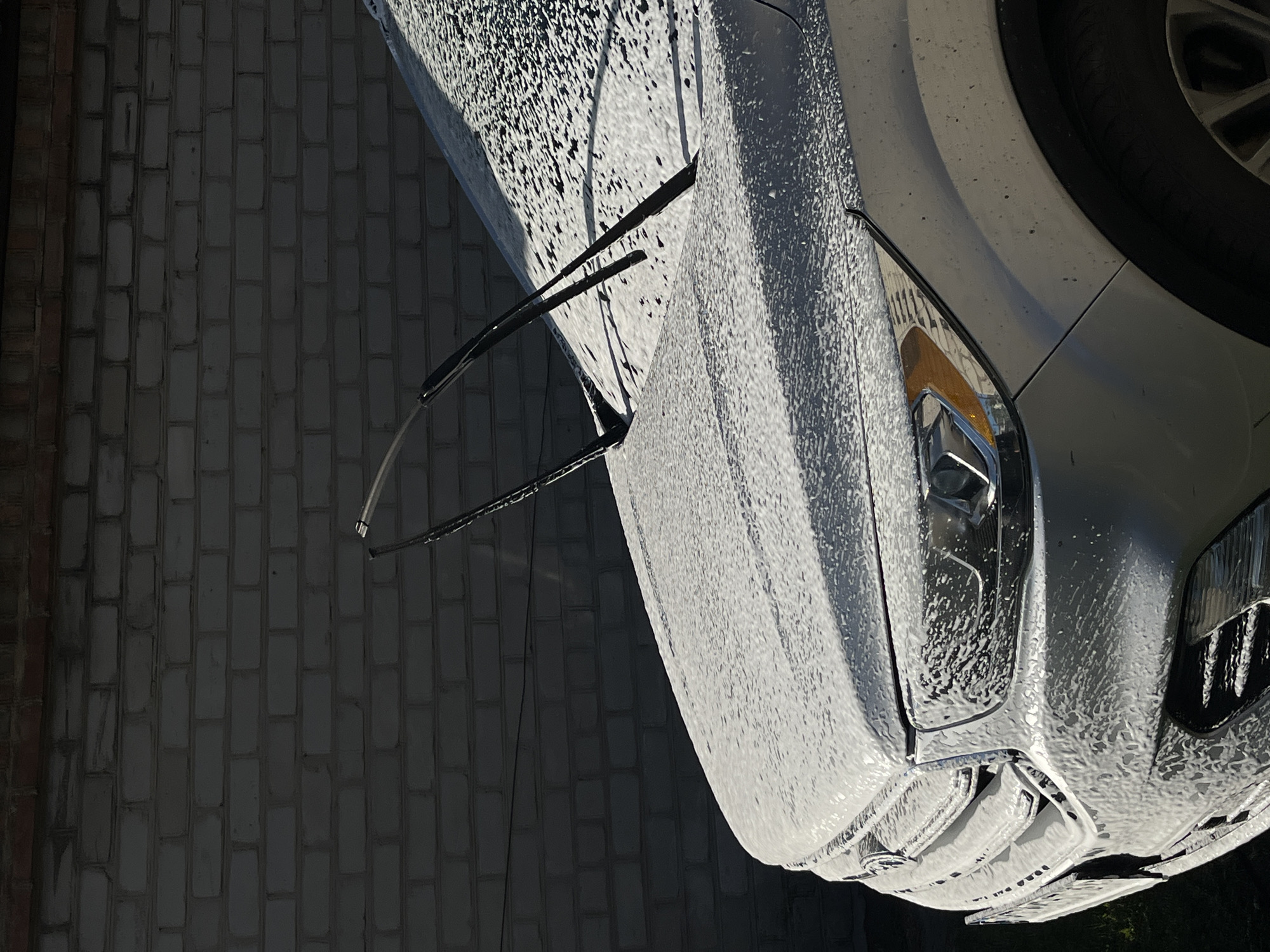 The image shows the front and hood of a white car covered in soap suds. The car is parked in front of a light grey brick wall. Two windshield wipers are visible on the hood, resting on the soapy surface. The front of the car, including the headlights and grille, is also covered in foam. Sunlight casts shadows on the car and the wall. The scene appears to be an outdoor car wash or detailing location.