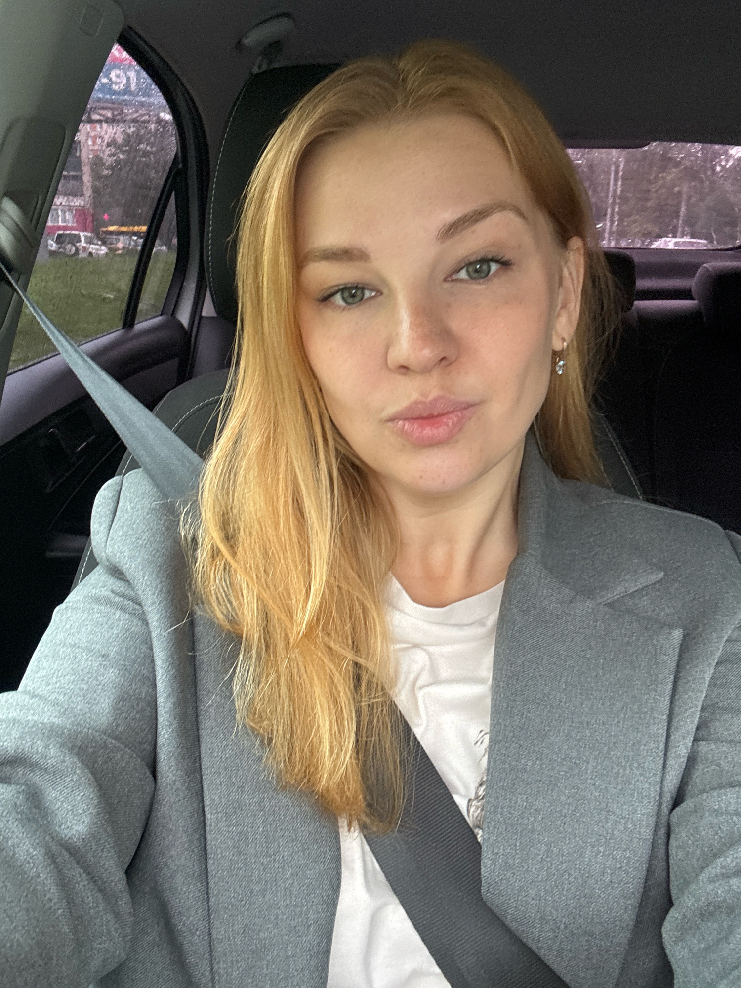 A blonde woman with fair skin and light green eyes is taking a selfie inside a car. She is wearing a grey blazer over a white t-shirt and a grey seatbelt is visible across her chest. Her blonde hair is shoulder-length and slightly wavy. She has freckles on her cheeks and her lips are slightly pursed. The background shows the interior of a car, with a window to her left revealing a blurry outdoor scene with buildings and trees. The woman is seated in the driver's seat. The car is likely parked or moving slowly in Rusanovo, Russia.