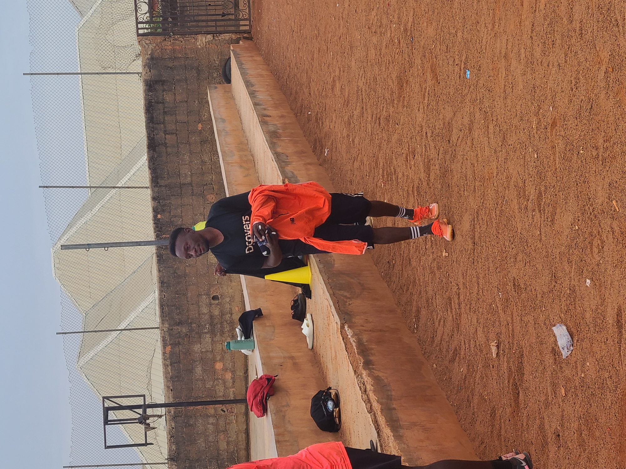 A medium shot depicts a black man wearing a black t-shirt and black shorts standing on a concrete structure. He holds an orange shirt. The location appears to be outdoors on a red clay ground with a background of a fence, a building with a white roof, and a basketball hoop. Several items, including shoes, bags, a water bottle, and a yellow cone, are placed on the concrete structure. The city and state are Unknown, Unknown.