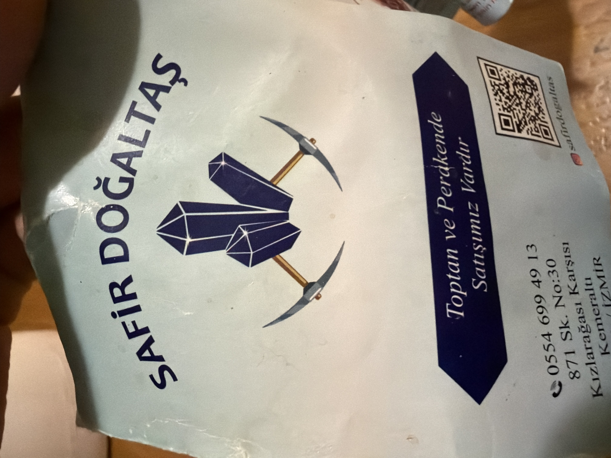 The image shows a business card for a company named "SAFİR DOĞALTAŞ". The company name is accompanied by a logo featuring stylized crystals and crossed pickaxes. A dark blue arrow shape contains the text "Toptan ve Perakende Satışımız Vardır," which translates to "We have Wholesale and Retail Sales." Below this is the company's contact information: a phone number (0554 699 49 13) and an address (871 Sk. No:30, Kızlarağası Karşısı, Kemeraltı, İZMİR). A QR code and an Instagram handle (@safirdogaltas) are also present. The background of the card is light blue/white.