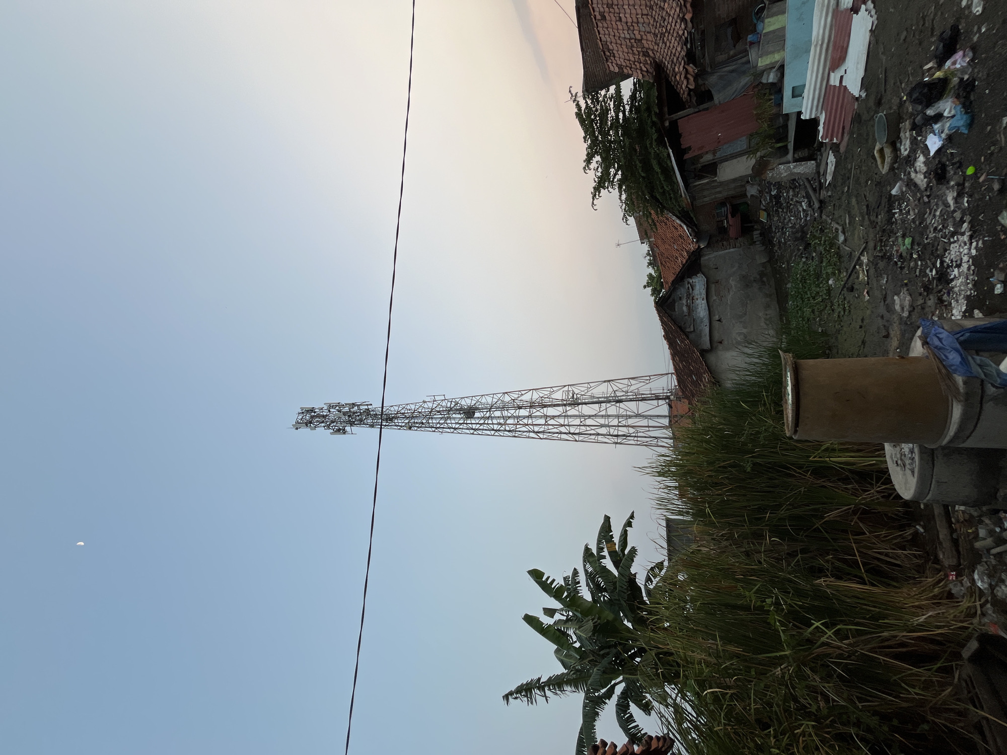 The image depicts a low-angle view of a tall, lattice telecommunications tower situated amidst a cluster of modest residential buildings and overgrown vegetation. The sky above is a pale blue, transitioning to a softer hue towards the horizon, with a sliver of the crescent moon visible. A power line stretches horizontally across the upper portion of the frame.
In the foreground, to the left, a dense patch of tall grass and banana trees is prominent. To the right of the vegetation, the telecommunications tower stands as the central vertical element, its structure intricate and metallic. Surrounding the base of the tower and extending towards the foreground are several circular concrete structures, possibly septic tanks or pipes, and a large, cylindrical object with a weathered surface, possibly a water tank or barrel. The ground in this area is littered with debris, including plastic bags, discarded materials, and what appears to be muddy ground with some standing water.
Behind the vegetation and debris, a series of low-rise buildings with tiled roofs are visible. Some of these structures appear weathered, with exposed brickwork and rudimentary building materials. The architectural style suggests a residential area. To the right of the telecommunications tower, more buildings and scattered debris are present. The overall impression is one of a somewhat neglected urban or semi-urban environment.
The image appears to have been taken in the late afternoon or early evening, given the soft lighting and the presence of the moon. Based on the context of the request, this scene is located in the city of Pekalongan, Indonesia.