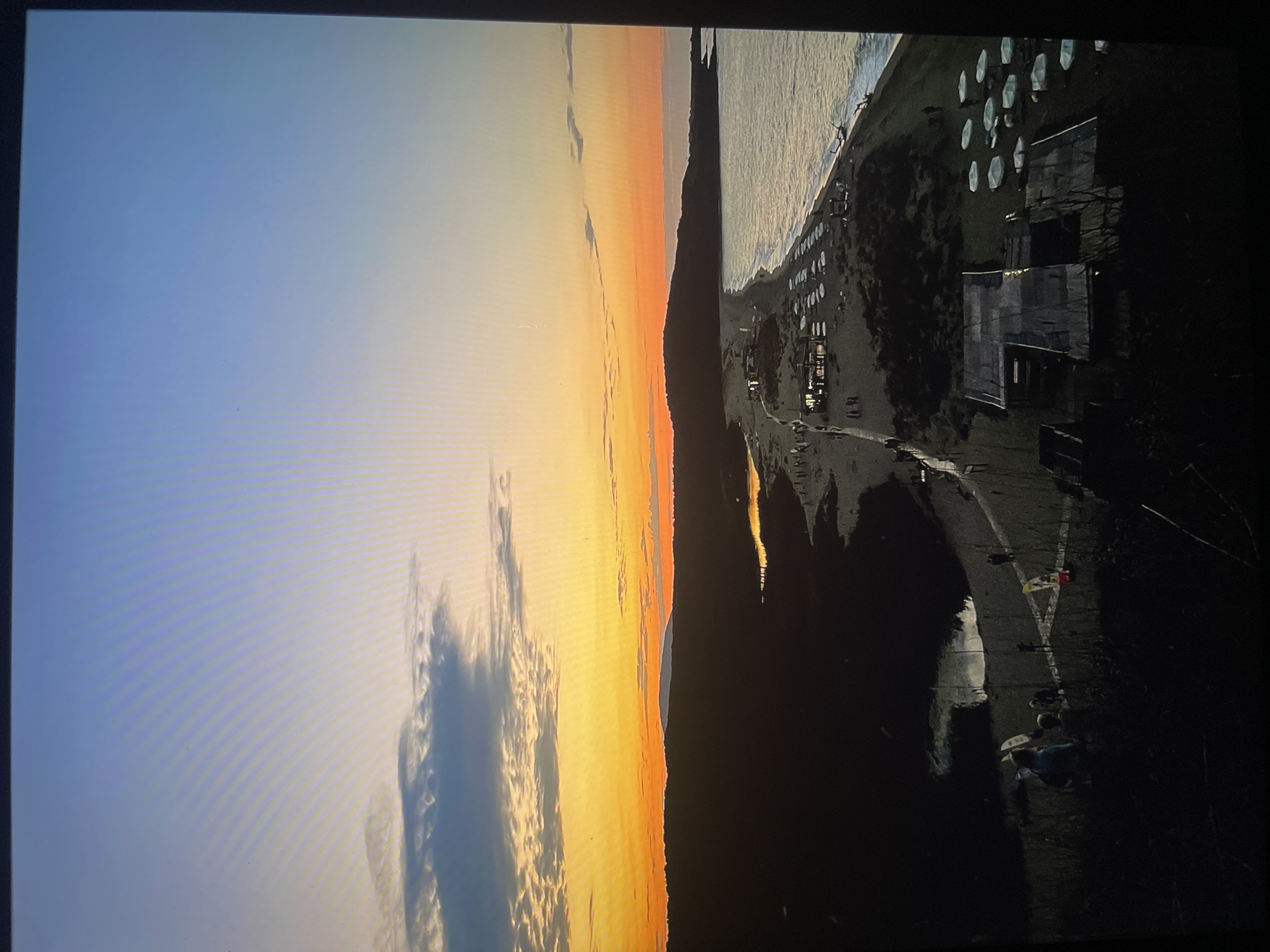 The image depicts a coastal scene at sunset. The sky transitions from light blue to orange, with some clouds present. Adjacent to the sky is a body of water. Parallel to the water is a stretch of land, which appears to be a beach or coastline, with structures and rows of objects visible on it. Dark shadows are cast across the land. People are seen moving on the ground. The location in the city of Unknown, Unknown is unknown.