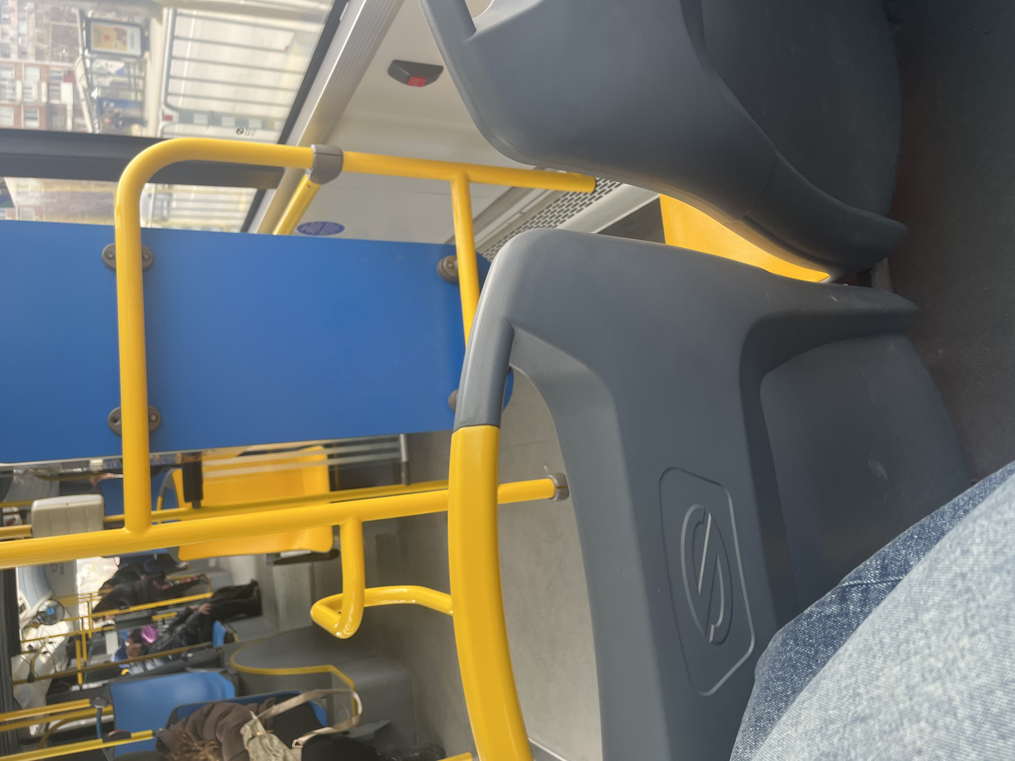The image depicts the interior of a public transit bus in Varna, Bulgaria, viewed from a seated passenger's perspective. The lower left quadrant of the frame shows the right knee and thigh of the photographer, dressed in light blue jeans. Two dark grey plastic bus seatbacks are prominent in the foreground, with a stylized 'S' emblem on the left seat.
The bus interior is characterized by numerous bright yellow metal handrails and support poles, some integrated with grey plastic connectors, extending throughout the cabin. Blue upholstered seats are visible on the left side of the bus, occupied by multiple blurred passengers. One passenger with brown curly hair, wearing a brown jacket, sits on the left, with a light-colored bag on their lap. Further back, another passenger in a dark jacket and blue or purple hat is seated, appearing to hold a pink object. A single yellow plastic seat is visible on the right side of the bus, behind the foreground grey seats. Blue wall panels are present on the left and center. The floor is predominantly light grey, transitioning to a textured black anti-slip surface near the windows. A red stop-request button is affixed to a white panel adjacent to the window.
Through the bus windows on the right, an urban scene is visible. A multi-lane street is separated from a sidewalk by a metal railing. A bus stop shelter with transparent walls and advertising panels stands on the sidewalk. In the background, several multi-story, light-colored residential or commercial buildings with numerous windows are visible under an overcast sky. Indistinct human figures are present near the bus stop. The scene captures a typical daytime moment within the city.