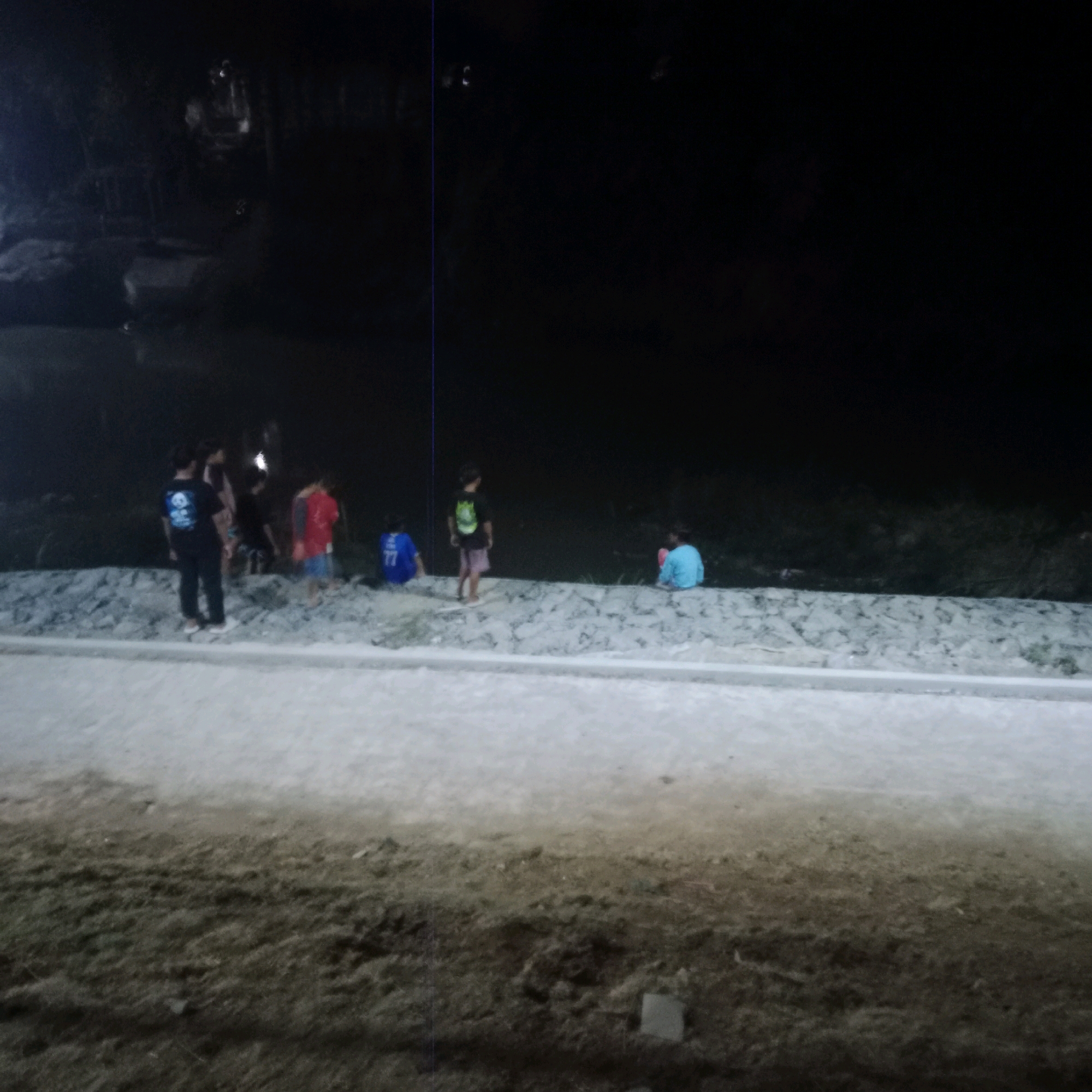 In Tlogorejo, Indonesia, at night, a group of five individuals, appearing to be children, are gathered by a body of water. One individual, dressed in a black t-shirt with a white design and black pants, stands facing left. To their right, another individual in a red shirt and light-colored shorts is partially visible. Further to the right, a third individual in a blue shirt with the number "77" is seated. The fourth individual, in a black t-shirt and light-colored shorts, stands facing away from the camera. The fifth individual, in a light blue shirt, is seated to the far right. The background is dark and indistinct, with the water's edge lined with rocks and a light-colored path or embankment in the foreground.