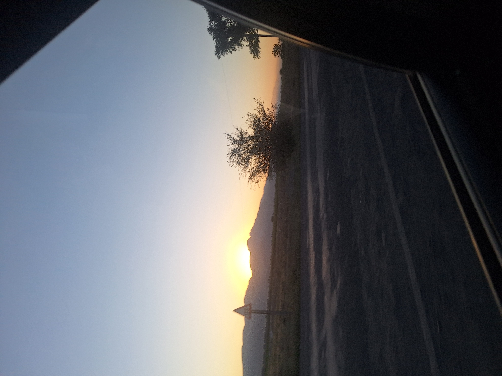 The image captures a serene sunset scene in the city of Chüy, Kyrgyzstan, viewed from the perspective of a vehicle. A golden sun is setting behind a range of low mountains, casting a warm glow across the horizon. In the foreground, a paved road is visible, with a traffic sign indicating a warning ahead. Dry grass and scattered trees dot the landscape between the road and the mountains. The sky transitions from a clear pale blue at the top to a soft yellow-orange near the horizon. The dark frame of a car window partially obscures the view on the right side of the image, suggesting the photograph was taken during a journey. The overall atmosphere is calm and tranquil, with the natural beauty of the Kyrgyz landscape taking center stage.