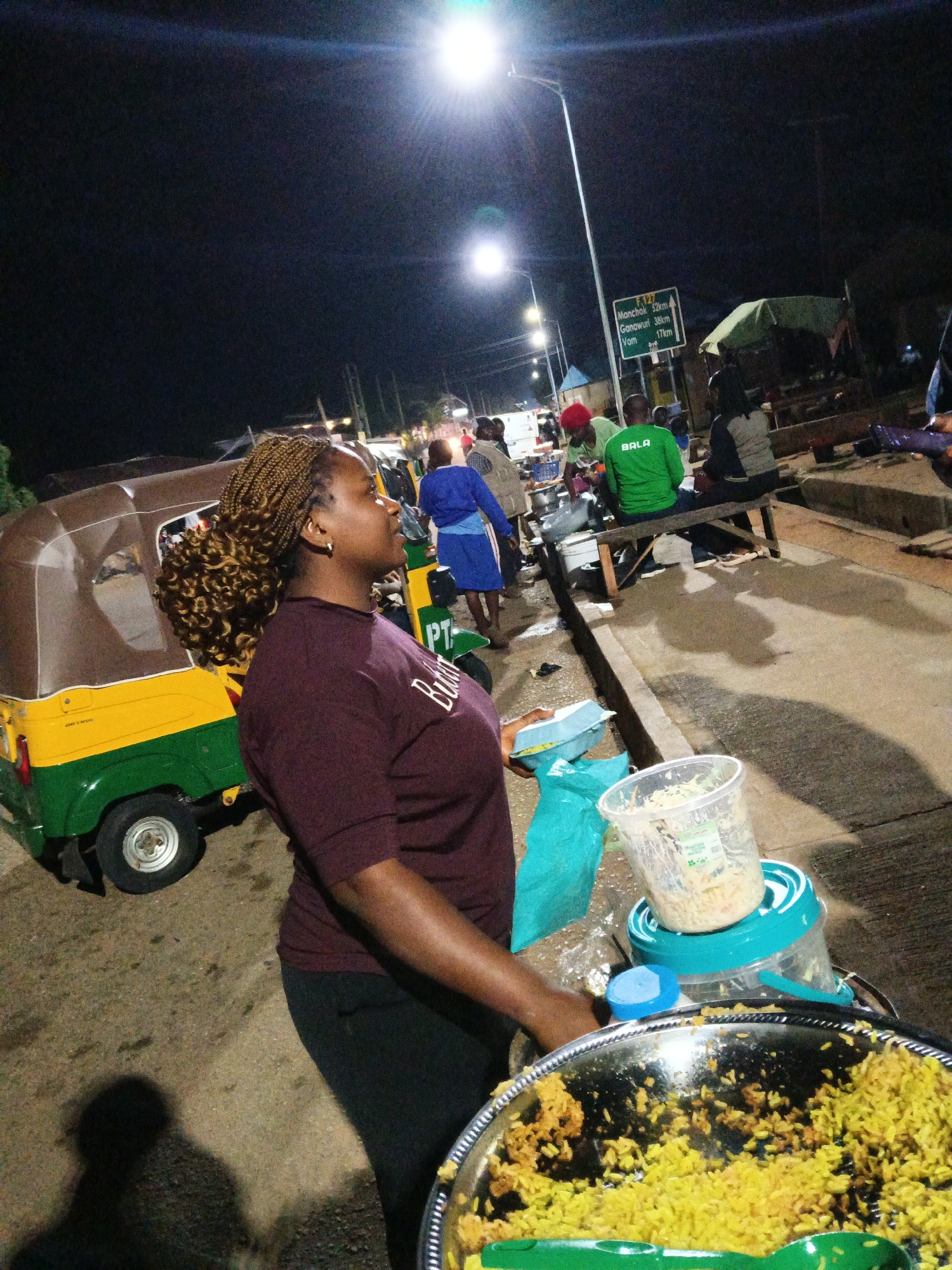 A street scene at night in Bukuru, Nigeria, captures a woman standing next to a food stall. She is wearing a maroon t-shirt and dark leggings, her hair styled in blonde braids pulled back. In front of her, a large metal platter is filled with yellow rice and a clear plastic container on a turquoise base holds a portion of what appears to be coleslaw. To her left, a yellow and green auto-rickshaw is parked.
Behind the woman, other individuals are visible. A man in a green long-sleeved shirt with "BALA" printed on it sits on a wooden bench with others. A signpost with white lettering on a green background is mounted on a pole in the background, indicating distances to Monchok (52km), Ganawuri (38km), and Vom (17km). The street is illuminated by streetlights, casting shadows on the concrete ground. The overall atmosphere suggests a bustling night market or roadside food vendor operation.