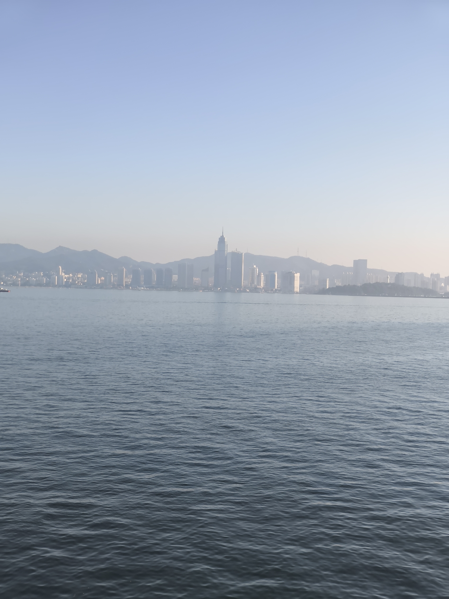 A wide shot captures a body of water in the foreground, with gentle ripples reflecting the hazy sky and a distant cityscape. The water appears calm, with subtle undulations. In the background, a dense skyline of buildings rises against a backdrop of rolling hills, partially obscured by a light mist. The architecture suggests a modern city, with various heights and styles of structures. A single red-hulled boat is visible in the far left of the image, moving across the water. The sky is a pale blue, with a gradient effect that lightens towards the horizon, indicating either early morning or late afternoon light. This scene is consistent with the coastal view of the city of Zhifu District, China, known for its port and urban development.