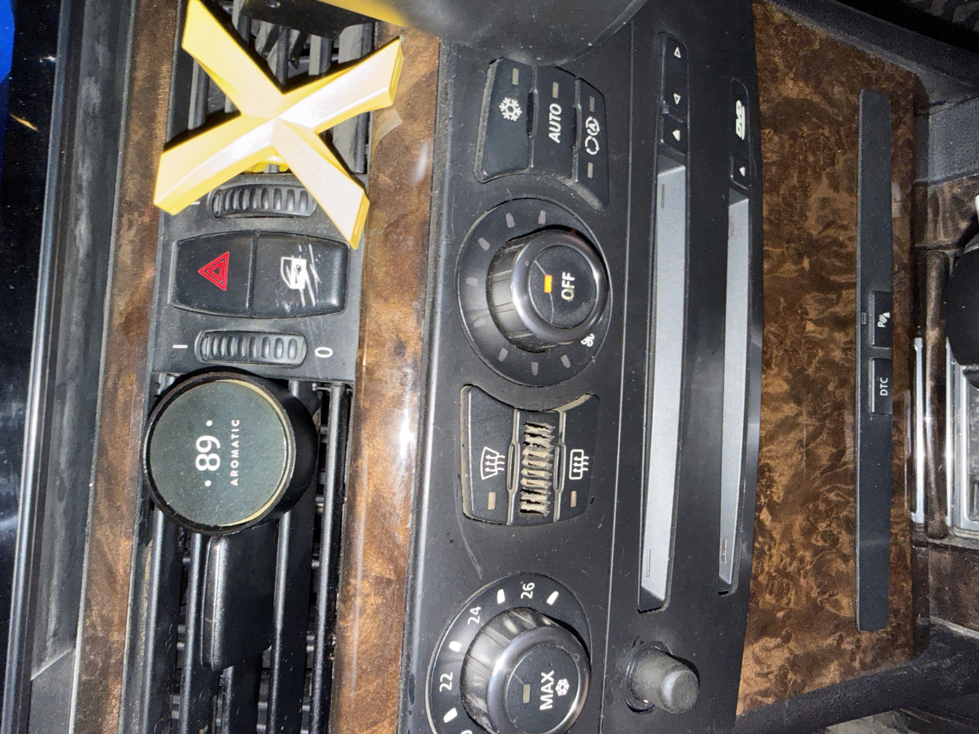 The image shows a close-up view of a car's dashboard. Visible elements include air vents, climate control knobs and buttons, a CD player slot, and trim with a wood-grain pattern. A round air freshener labeled "89 AROMATIC" is clipped to one of the air vents. A yellow plastic "X" shaped object is positioned on another air vent. The scene appears to be indoors, likely within a garage or parked car. There are no people visible. The context of Stara Zagora, Bulgaria, is not visually represented in the image itself.