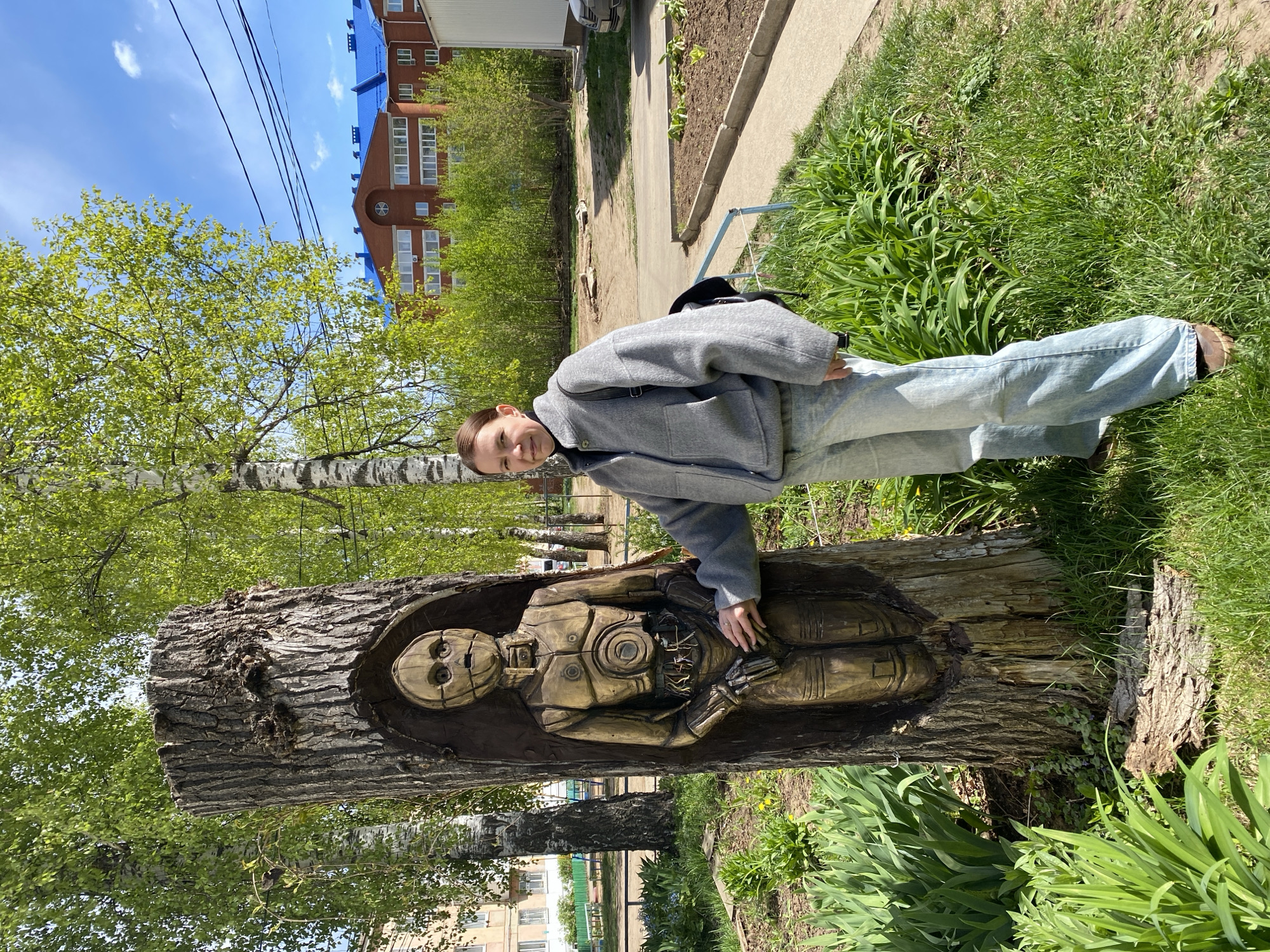 A woman stands next to a tree trunk carving in what is likely the city of Chaikovsky, Russia. The carving depicts a humanoid robot-like figure. The woman is wearing a light gray jacket, light blue jeans, and brown shoes. She is holding the carving. The carving is of a metallic color. The surrounding environment consists of grass, flowers, other trees, and a building. Power lines are visible in the background. The sky is partially covered by light clouds.