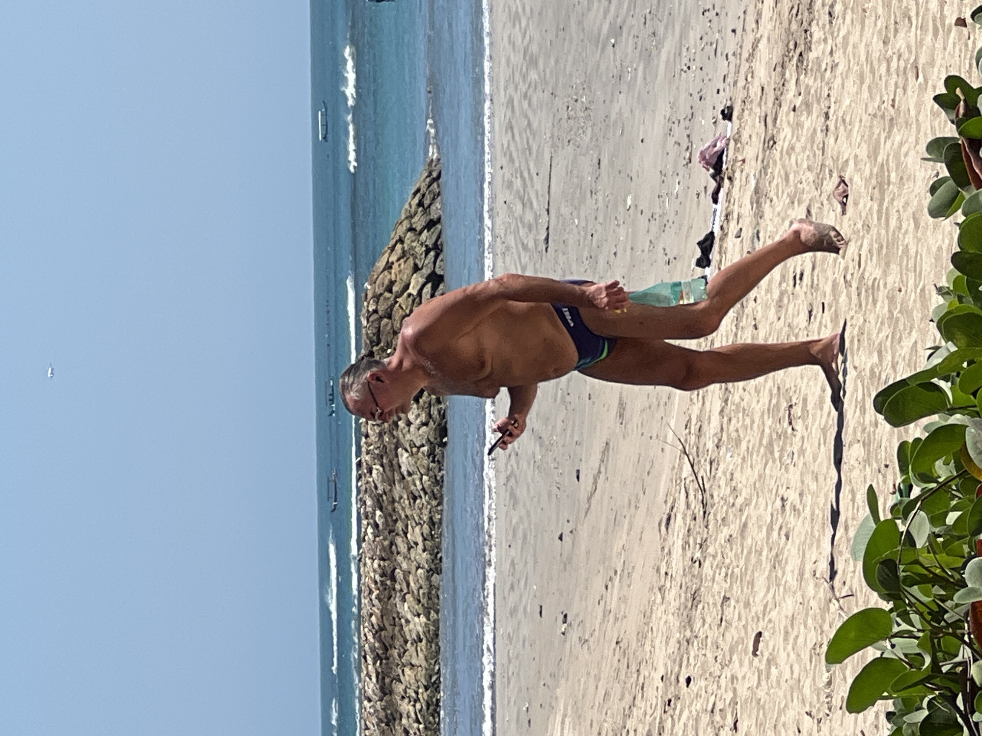 A middle-aged man with glasses and graying hair walks along a sandy beach. He is wearing dark swim trunks with a green stripe. In his right hand, he holds a smartphone, and in his left hand, he carries a clear plastic bottle. The man is walking towards the right of the frame. In the background, the ocean is visible with moderate waves breaking on a stone barrier that extends into the water. Several boats are anchored in the distance. The sky is clear and blue. In the foreground, lush green leaves partially obscure the bottom of the frame. This scene is consistent with the beaches in Kuta, Indonesia, known for their wide sandy shores and ocean access.